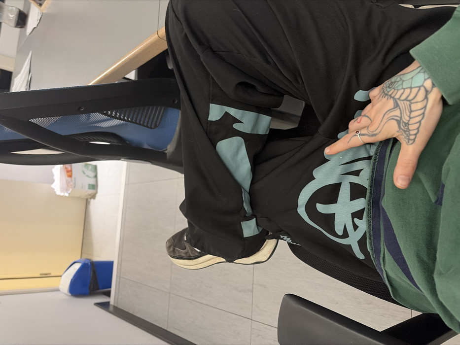 The image presents a first-person, downward-angled view of an individual's legs and left hand within an indoor office or commercial setting. The person wears black athletic pants featuring light blue graphic designs on the thighs, a dark green long-sleeved top, and black sneakers with thick, off-white soles. The visible left hand has a distinct light blue tattoo depicting a serpent or dragon's head and neck, and a silver ring with a red stone is present on the index finger. The individual is seated in an office chair characterized by a black frame and a blue mesh backrest.
The background reveals a minimalist office environment. To the right, a dark-colored table holds scattered papers and a dark glass bottle. The floor is covered with light-toned rectangular tiles. In the mid-ground, a blue and white trash can with a black liner is positioned against a light-colored wall, adjacent to a closed light-colored door. A stack of white paper or boxes is also visible on the floor to the right of the trash can. No specific identifying features are present in the image to determine a precise location within the city of Sofia, Bulgaria.