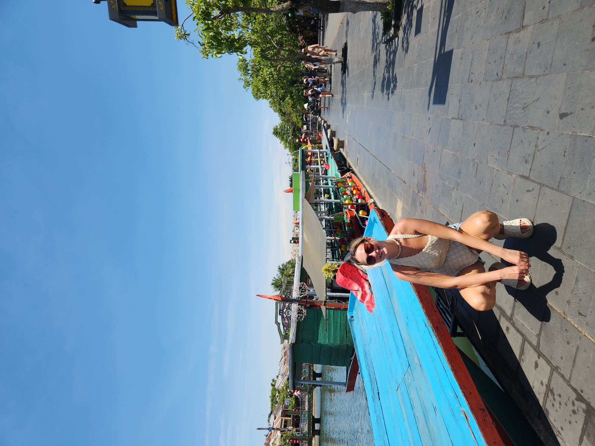 In Hội An Ward, Vietnam, a woman with blonde hair and sunglasses is squatting on a paved walkway next to a canal. She is wearing a white crocheted top and denim shorts, and she is smiling at the camera. The walkway is lined with trees and a row of boats docked along the canal. The boats are brightly colored and decorated with lanterns. In the background, there are buildings with traditional Vietnamese architecture. The sky is clear and blue, and the sun is shining.