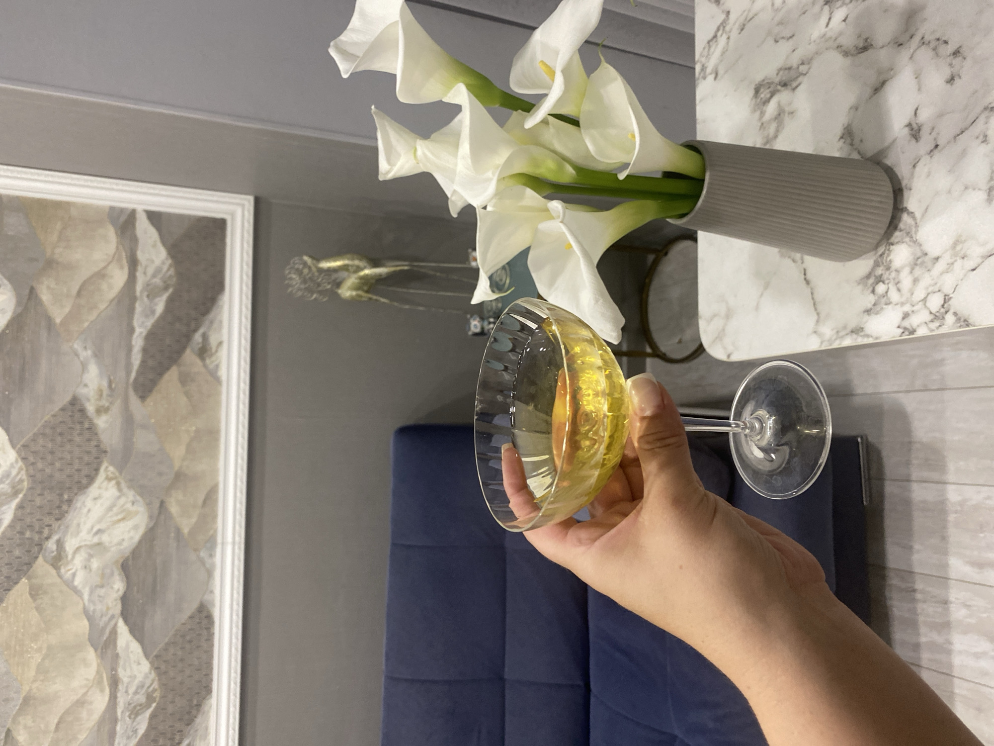 The image depicts an indoor scene in the city of Sochi, Russia, with a first-person perspective focused on a hand holding a drink. In the immediate foreground, a person's left hand, with light-colored manicured fingernails, grasps the stem of a clear, coupe-style glass containing a transparent yellowish liquid. To the right, a bouquet of approximately eight white calla lilies with yellow spadices is arranged in a ribbed, light-gray ceramic vase. The vase and a portion of the glass are situated on a table with a white surface featuring gray marble-like veining. In the mid-ground, a dark blue, tufted sofa is visible to the left. The wall behind the sofa and table is painted a solid gray. Above the sofa, a large, rectangular framed artwork is mounted, depicting an abstract, layered landscape or mountain range in shades of beige, gray, and white, framed in white. Further to the right in the background, a golden-colored, slender, abstract sculpture resembling a stylized animal stands on a surface. The floor beneath consists of light-colored wooden or laminate planks.