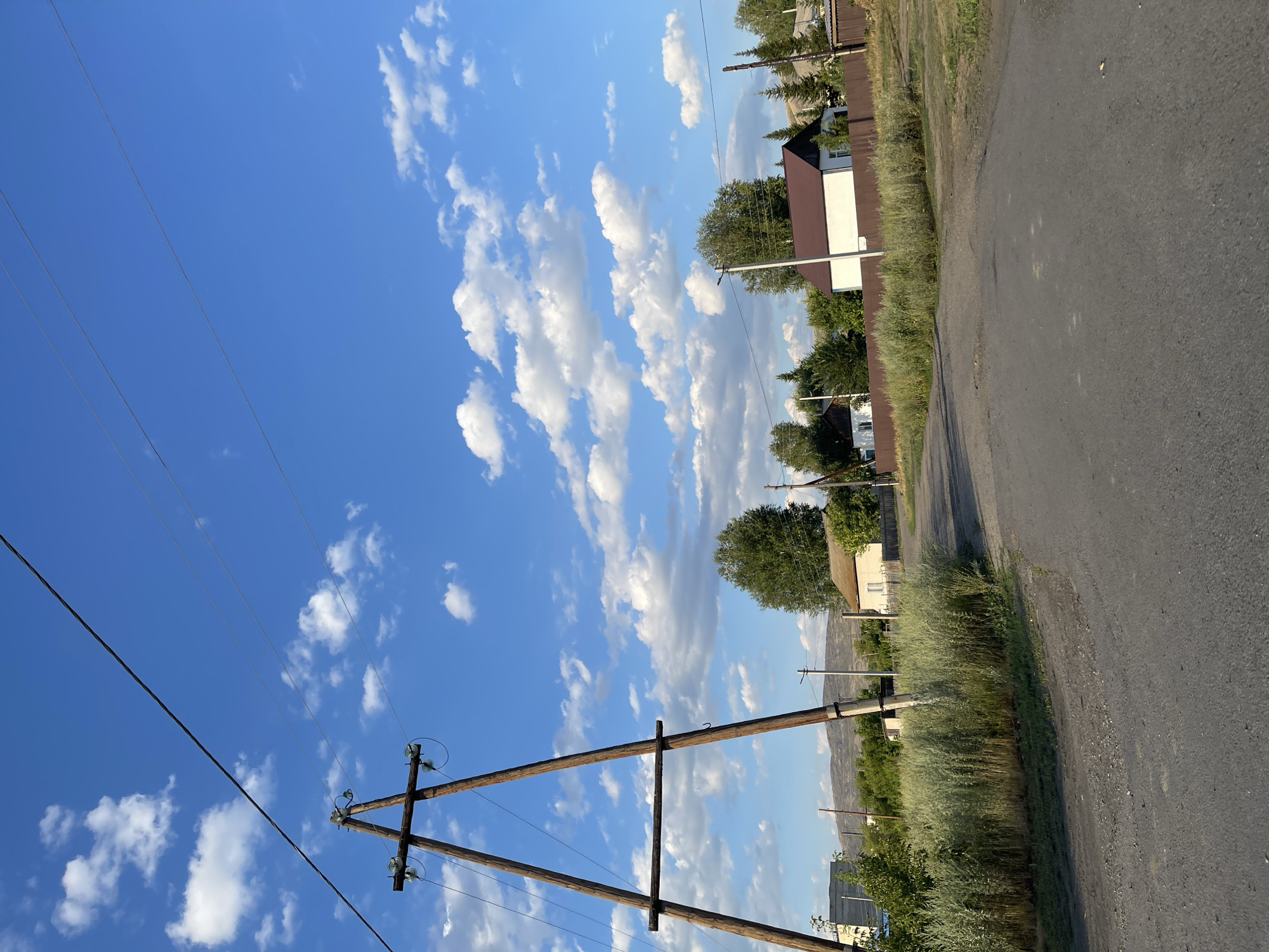 The image shows a rural street scene in Predgornoe, Kazakhstan, under a bright blue sky with scattered white clouds. A wooden power line pole, constructed from two angled posts joined by a horizontal beam, stands prominently on the left side of the road. Power lines stretch across the upper portion of the frame.
The road itself is unpaved and appears to be a dirt or gravel track, with some patches of asphalt visible. To the right of the road, a row of houses with dark brown roofs and white walls are visible, separated by a brown wooden fence. Green trees and tall, dry grass are abundant throughout the scene. In the background, hills or mountains can be seen in the distance. The overall impression is one of a quiet, rural settlement.