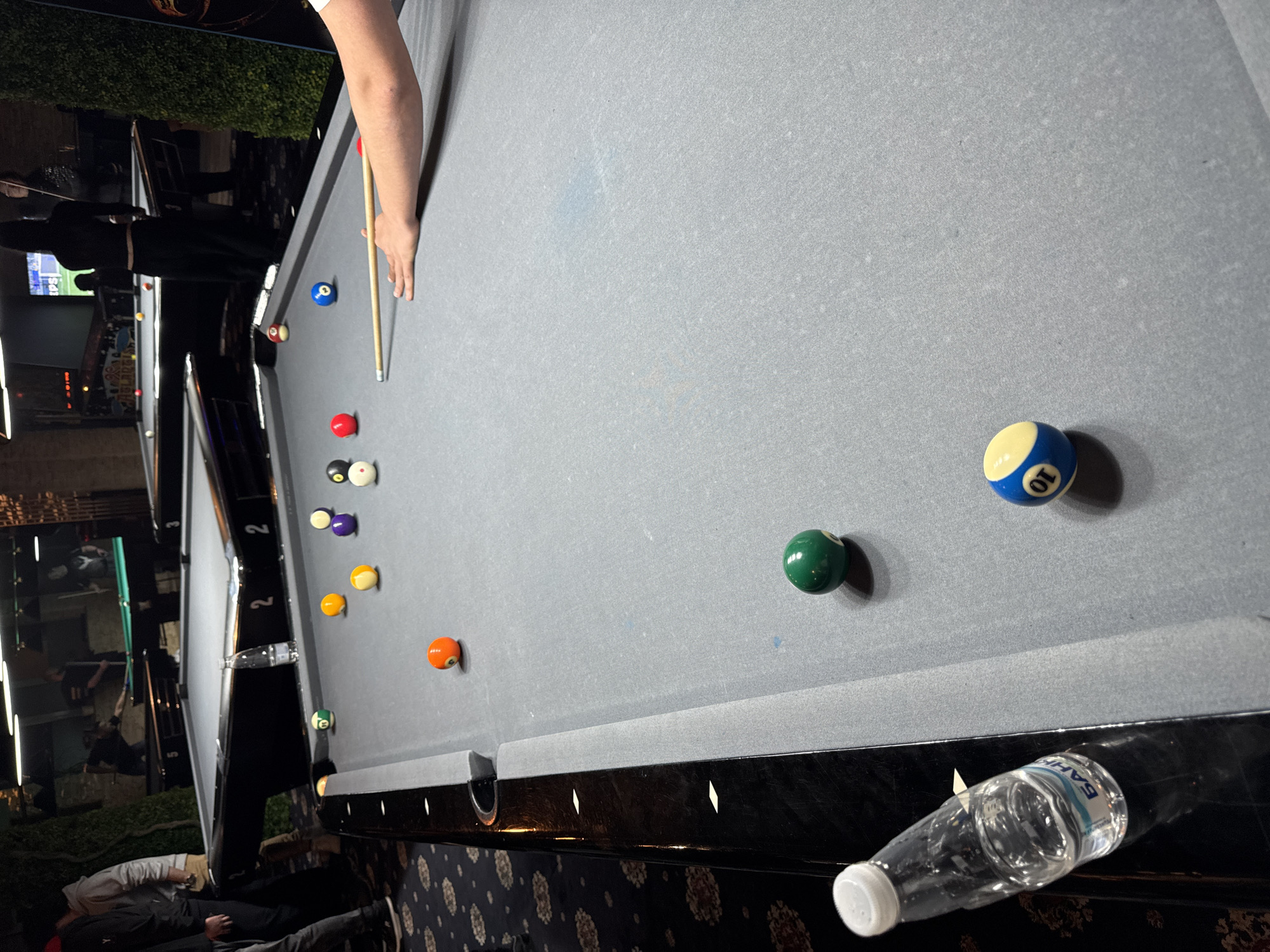 A medium-angle, rotated, indoor shot depicts a pool table with several billiard balls scattered across the grey felt surface. A person's arm holds a cue stick, positioned near the cue ball and red ball. In the foreground, a partially empty plastic water bottle lies on the table’s edge. The balls visible include a striped 10-ball, a green ball, an orange ball, two yellow balls, a purple ball, a red ball, and two balls with white coloration. Background context reveals another pool table adjacent to the primary one. The setting appears to be a bar or recreational area, identifiable by ambient lighting, and patrons visible in the periphery. The location is unknown, but the user requests the location is in Plovdiv, Bulgaria.