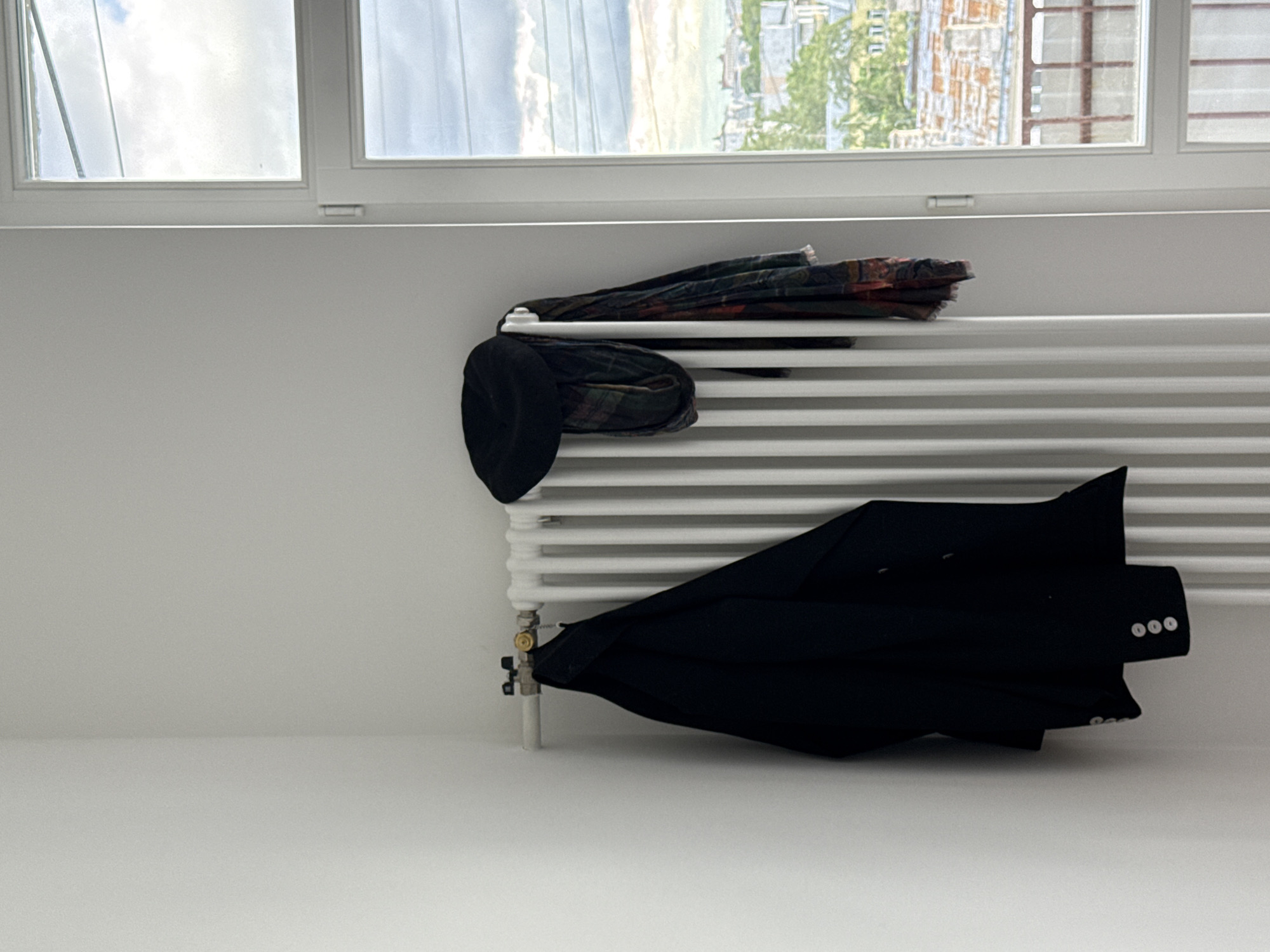 The scene depicts a white wall with a white radiator beneath a window. A black hat, a tartan scarf, and a black jacket are draped over the radiator. The view from the window includes buildings, trees, and a cloudy sky. The location cannot be determined.
