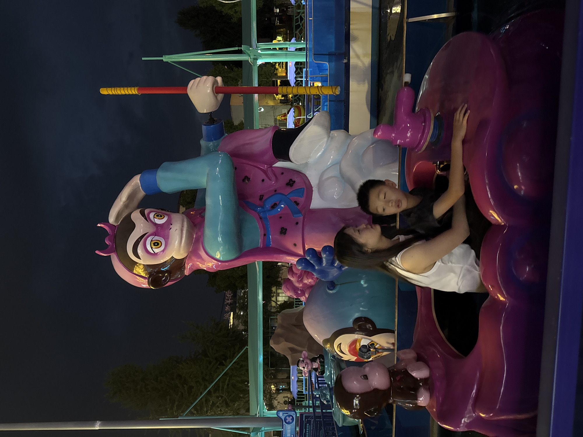 In Yiwu, China, a nighttime amusement park scene features a vibrant, oversized cartoonish sculpture of Sun Wukong, the Monkey King, holding a red and yellow staff. Below the sculpture, a mother and son are seated in a boat-like amusement ride, which is a deep purple color with swirling, organic shapes. The mother, wearing a white sleeveless top, looks to her right, while the son, in a dark t-shirt, gazes forward with his mouth slightly open. Another smaller monkey sculpture is visible in the boat. The ride appears to be in a pool of water, with reflections visible on the surface. The background is dark, indicating nighttime, with some trees and structures faintly illuminated. The overall impression is one of lively entertainment and family fun.