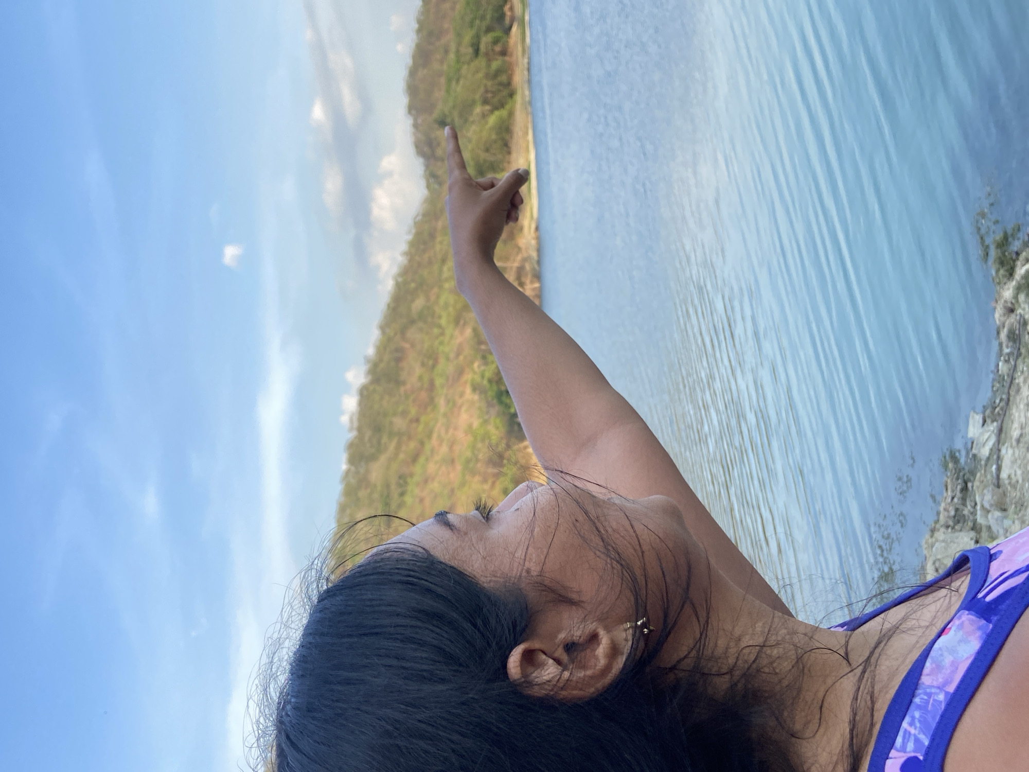 A woman with dark hair is pointing towards a hilly, forested area across a body of water with a blue hue under a blue sky. The location appears to be outdoors, possibly near a lake or reservoir in the Tinaquillo, Venezuela, area. The woman is wearing a blue and purple top. Her arm is extended, pointing at the landscape.