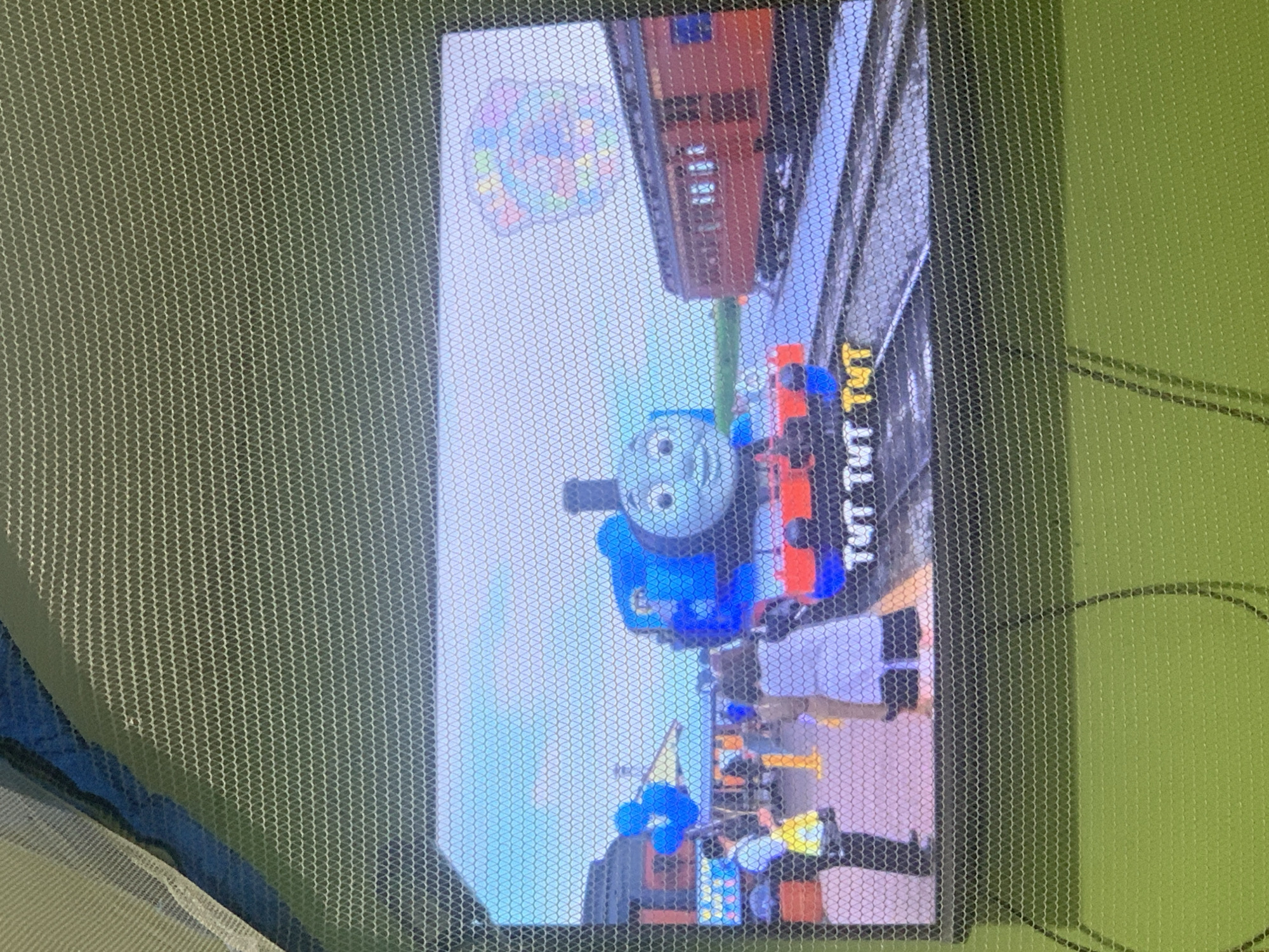 The image displays a screen, likely a television or monitor, displaying a scene from the children's show "Thomas & Friends." A blue cartoon train, identified as Thomas, is prominently featured on a railway track. To the right of Thomas is a red train car. In the background, there are several figures standing on a platform near a building and some blue balloons. Text on the screen reads "TUT TUT TUT". The screen itself is covered by a mesh netting, suggesting it is either outdoors or behind a protective barrier. The background behind the screen appears to be a green surface, possibly grass or fabric. There is no visual information within the image to suggest a location in Palembang, Indonesia.