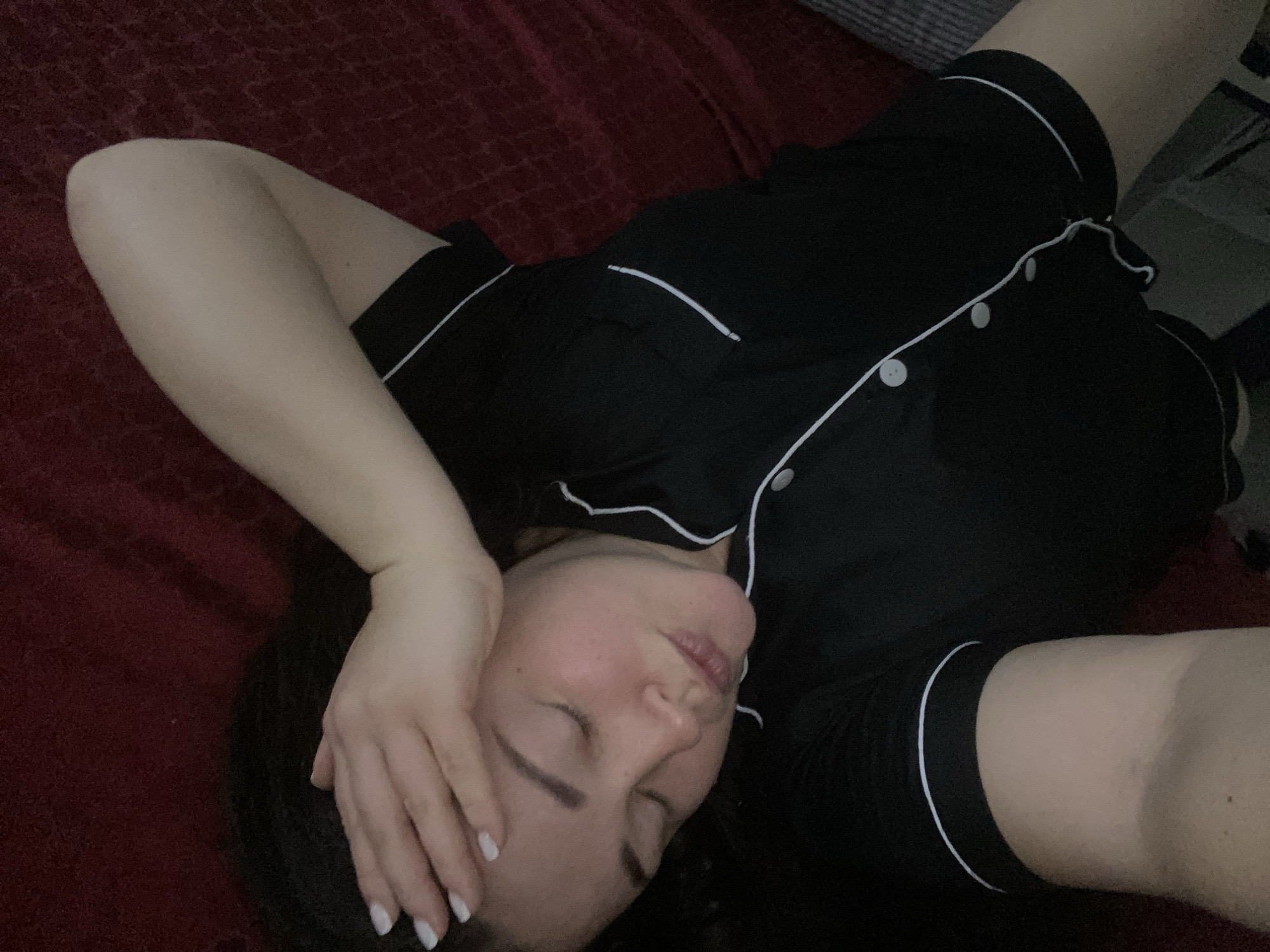 A woman with long dark hair lies on a red bed, wearing black pajamas with white piping. Her eyes are closed, and one hand rests on her forehead. The photo seems to have been taken indoors, possibly in a bedroom, but there is no evidence to determine a location in Sochi, Russia.