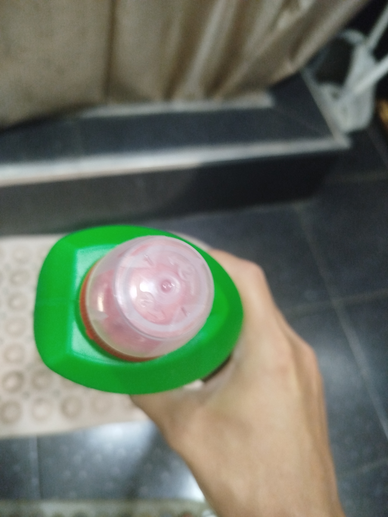 A close-up, overhead shot shows a hand holding a green plastic bottle with a pink, translucent cap. The bottle appears to be a cleaning product. The background is out of focus, but hints of a tiled floor, a patterned mat, and a draped fabric can be discerned. The scene is indoors, possibly a domestic setting. The image does not contain enough visual information to determine its specific location within Batumi, Georgia.