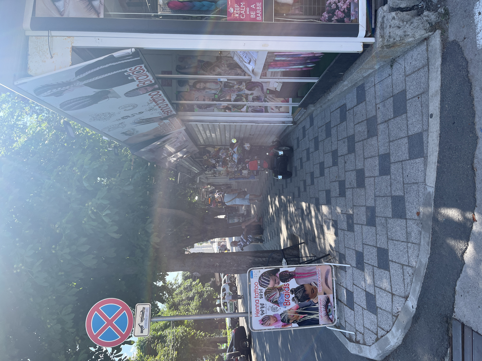 In Obzor, Bulgaria, a street scene unfolds under the dappled sunlight filtering through trees. A sidewalk, paved with grey and dark grey square tiles, runs alongside a row of small shops. In the foreground, a white A-frame sign displays advertisements for "Henna tattoo," "Braids," "Lash lamination," and "Brows lamination," featuring images of braided hairstyles and close-ups of eyes. To the left of the sign, a traffic sign prohibits stopping and parking, with an additional panel indicating towing and a phone number.
Further down the sidewalk, a red and black mobility scooter is parked. The shops appear to be selling hair extensions and braiding services, with colorful braids visible in display windows. One shop has a large banner advertising "Henna Tattoo" and "Braids" with images of braided hair. Another shop window displays a poster that reads "KEEP CALM AND BE A BARBIE."
Several people are present in the scene. A man in a striped shirt sits on a stool under a tree, looking towards the shops. Further down the street, a woman in a light-colored dress and a straw hat walks away from the camera. In the background, a white SUV with "SOD" written on it is parked. The overall atmosphere suggests a commercial street in a tourist area. The sunlight casts strong shadows on the pavement.