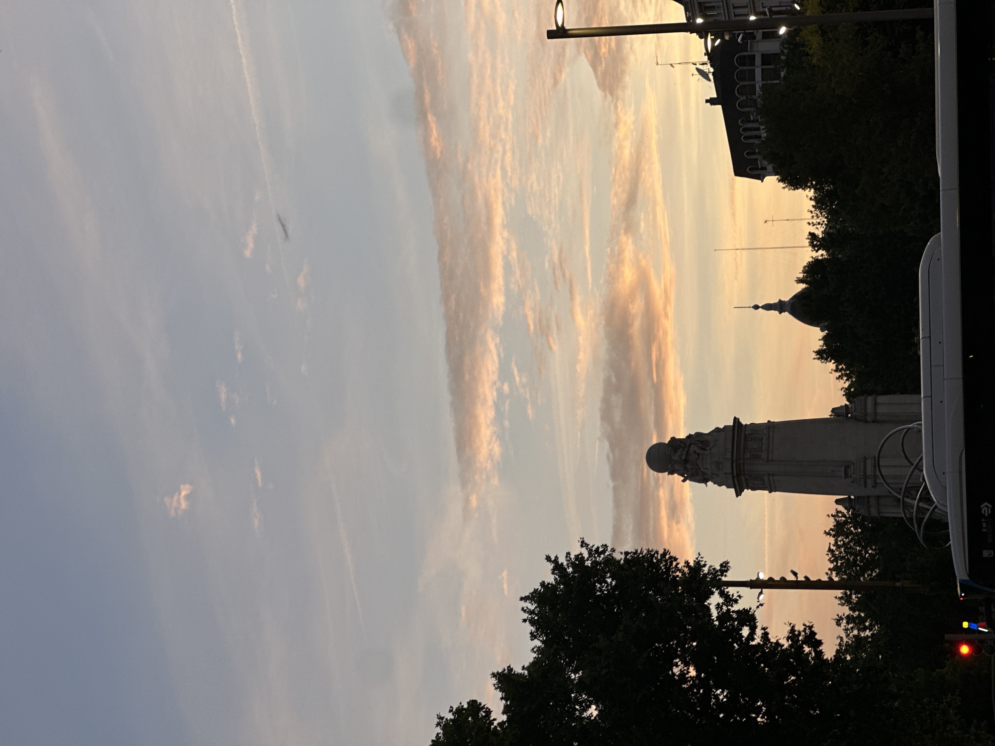 The image depicts a vertical view of the sky and surrounding cityscape during a sunset in Madrid, Spain. The sky is filled with striated clouds tinted with orange and gold hues of the setting sun. The foreground includes a monument, identified as the Monument to Cervantes in Plaza de España, Madrid. Silhouetted trees line the bottom of the frame and surround the monument. A street lamp is partially visible, and a bus is present on the right edge of the frame. The light is generally dim, indicating twilight.