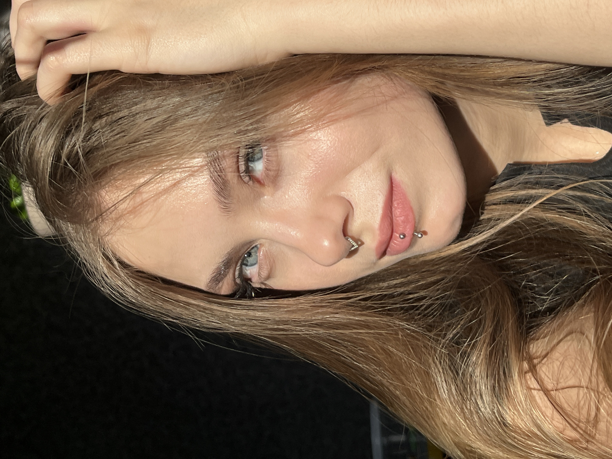 A young woman with light brown hair and blue eyes is shown from a close, slightly angled perspective. She has facial piercings on her nose and lip. The background is dark. No information is available about whether this image was taken in the city of Фоки, Russia.