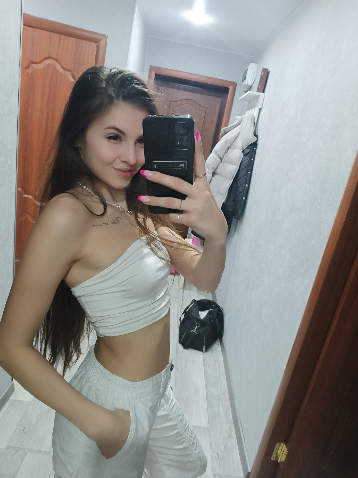 A young woman stands in a hallway or foyer, taking a selfie in a mirror. She is wearing a white one-shoulder top and white pants. Her left hand is in her pocket, and her right hand holds a black smartphone with a pink nail polish on her fingernails. She has long brown hair and a necklace. A black bag is on the floor near the wall. The walls are light gray. Two wooden doors are visible. Coats hang on a white shelf on the wall. The location is a hallway in a residence located in the city of Chaikovsky, Russia.