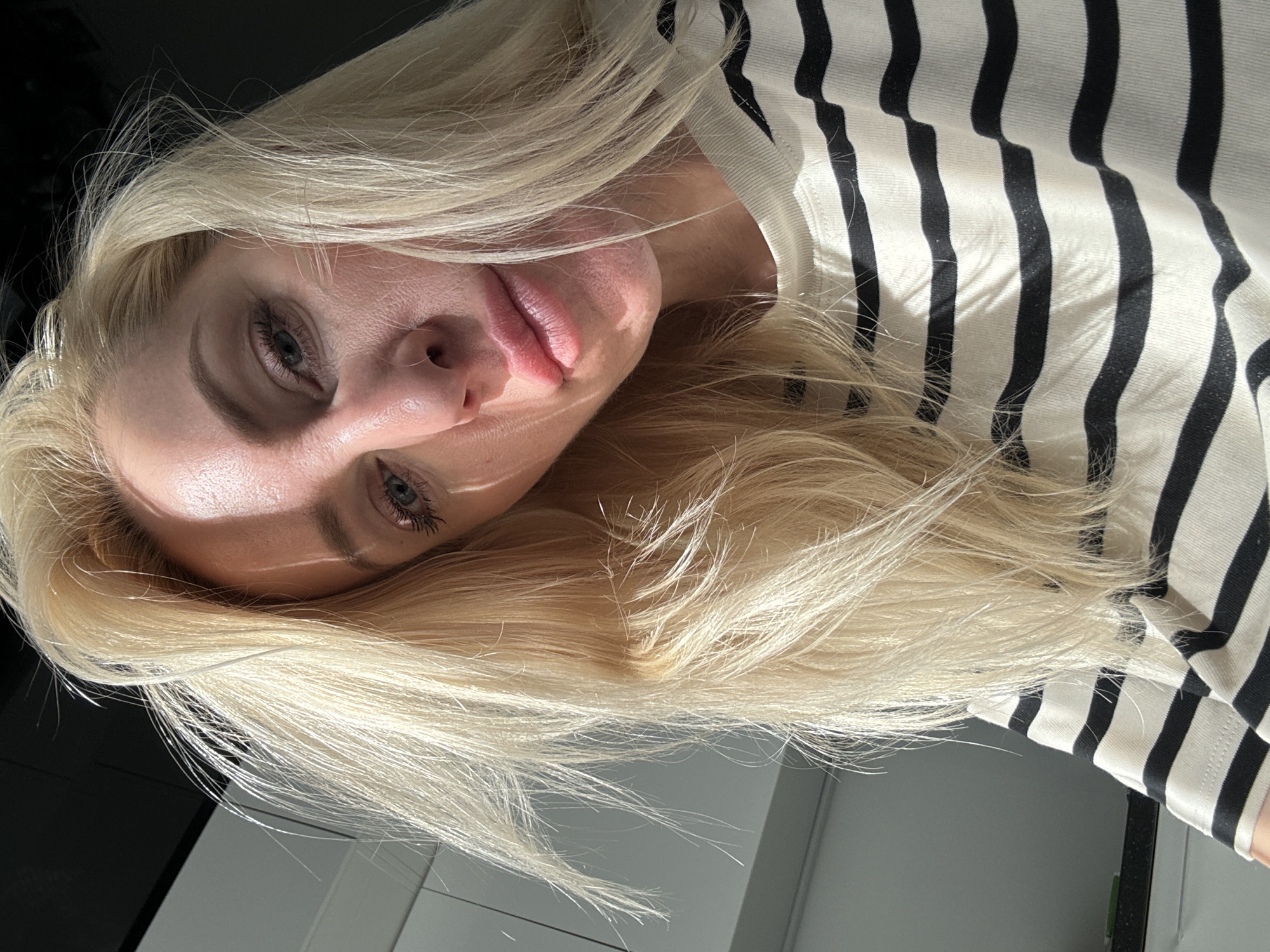 An extreme close-up selfie shows the upper body of a fair-skinned blonde woman wearing a black and white striped shirt. The woman is looking down and to her left, with her blonde hair framing her face and falling across her shoulders. Sunlight casts shadows across her face, highlighting her features. The background is out of focus, but appears to be an indoor setting with white cabinetry and a dark countertop. The overall lighting suggests natural light coming from a window. The image is high resolution, with sharp details on the woman's skin and hair.