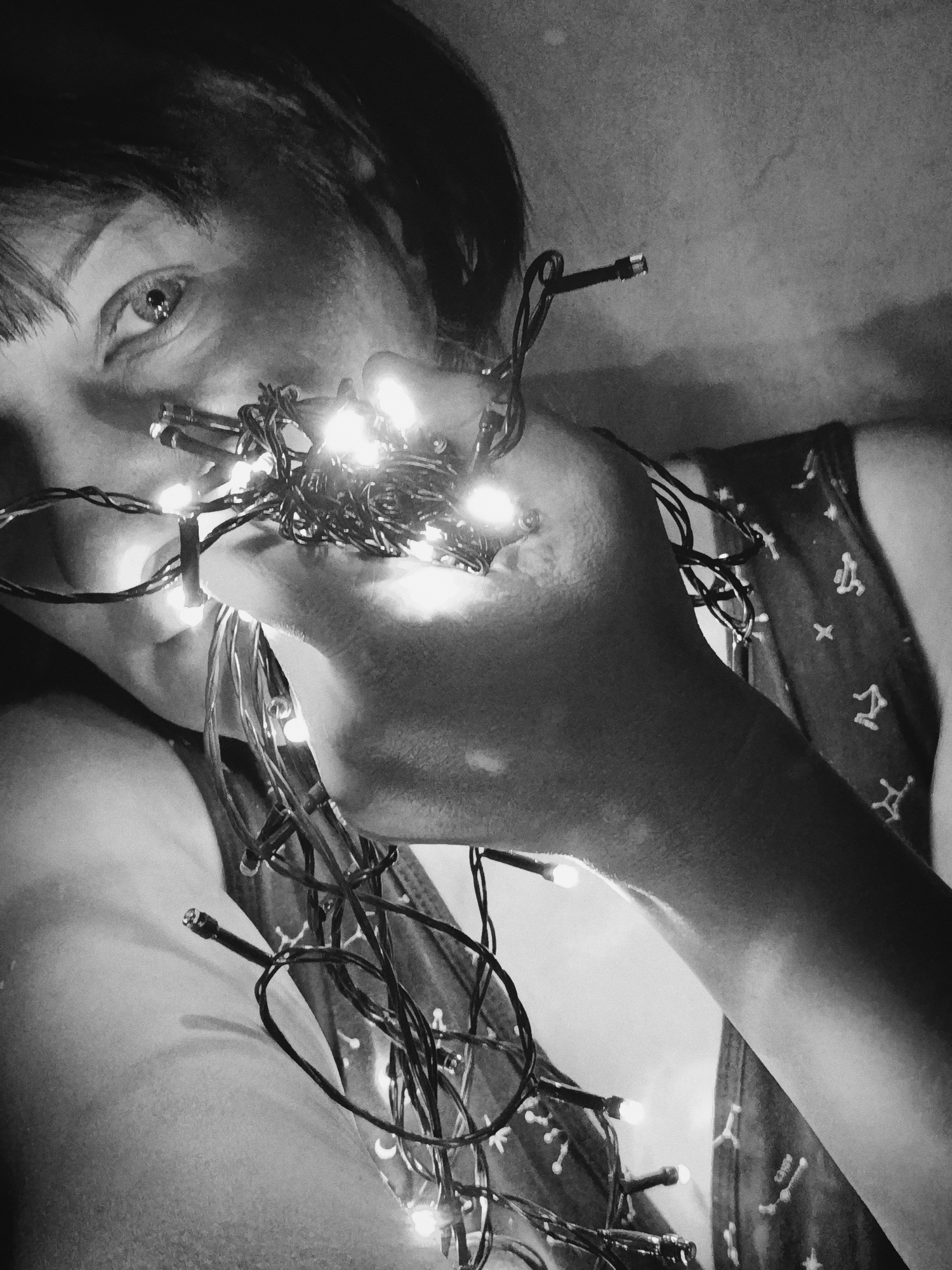 A black and white, close-up shot depicts a person holding a string of lit Christmas lights. The person, presumably female, has short hair with bangs and is looking directly at the camera with one visible eye. The string of lights is wrapped around the person's hand and held near their face, with several lights illuminated. The person is wearing a patterned tank top. The background is a blurred, neutral tone. The location and city are unknown.