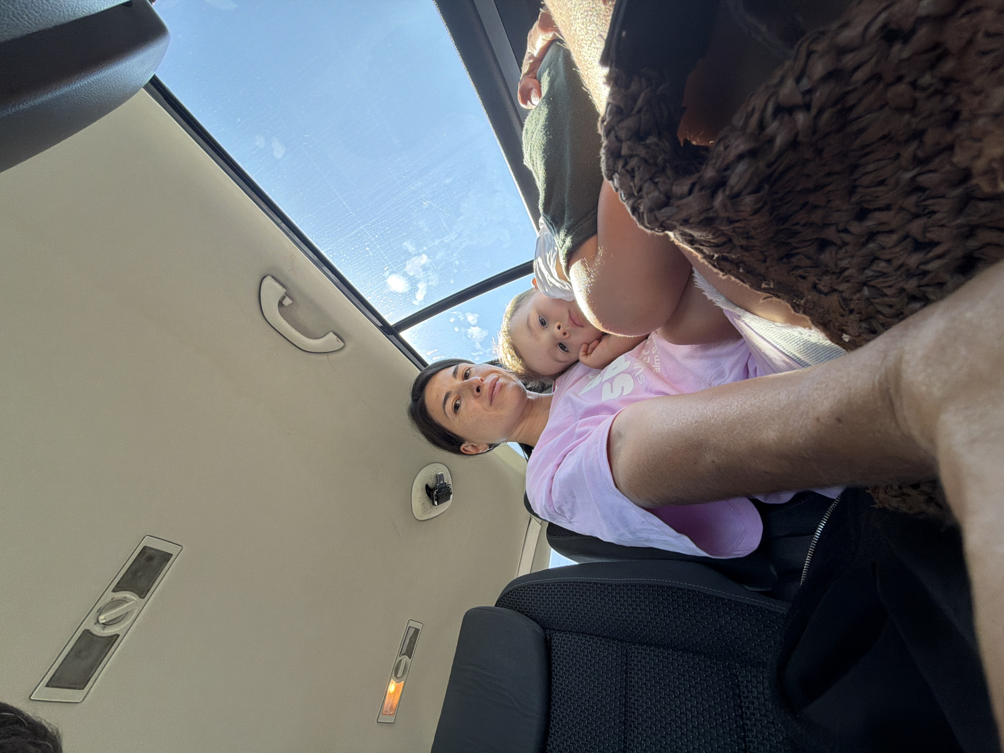 The image shows a woman in a car with a young child. The woman is wearing a pink t-shirt and the child is wearing green shorts. They are both looking at the camera. The car appears to be moving as evidenced by the blur of the scenery outside the window. The sky is blue and there are some clouds. The interior of the car is visible, including the ceiling and seats. The scene is a typical everyday moment captured in the city of Kruševica, Montenegro, likely during a road trip.