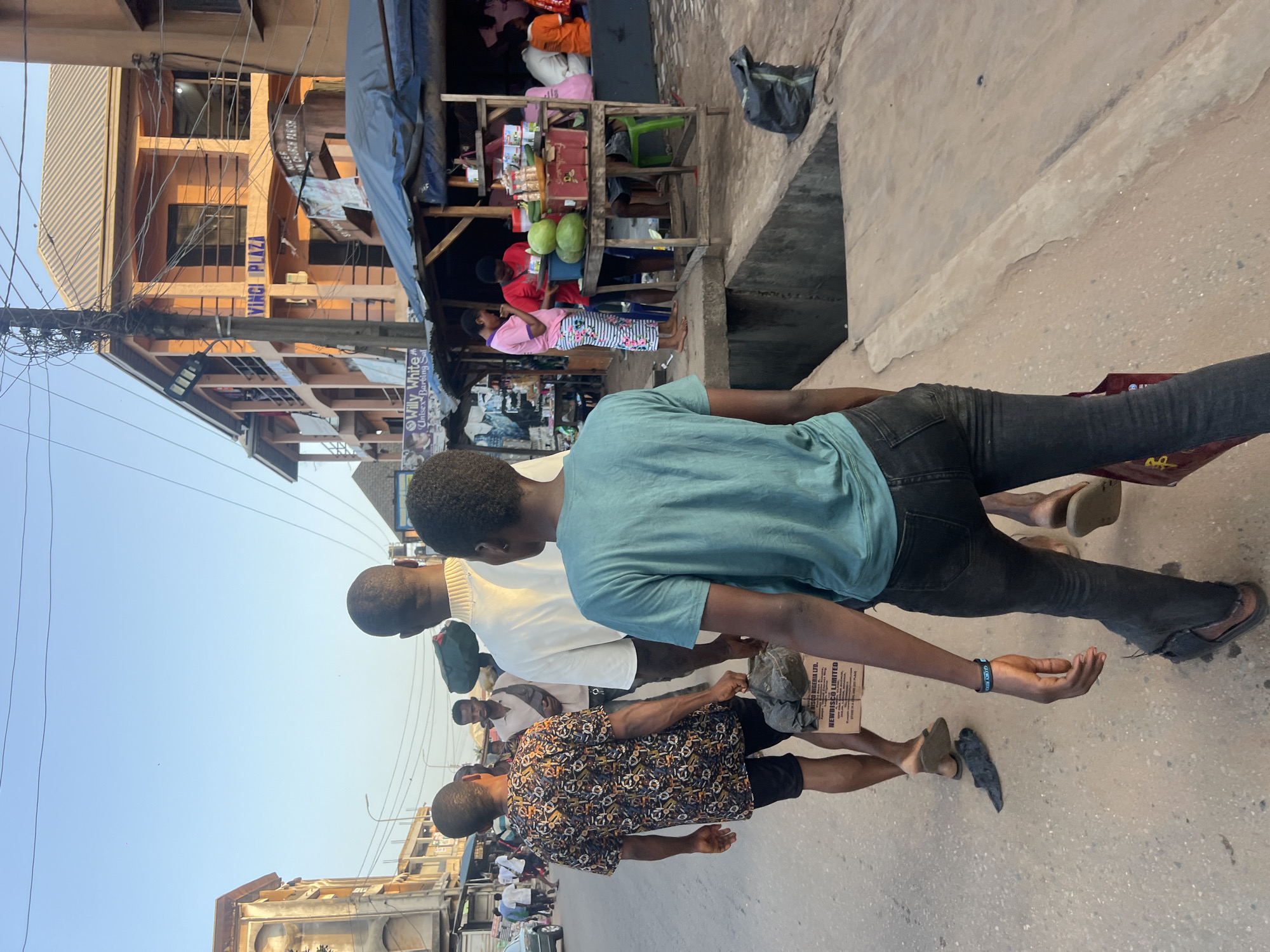 The image captures a street scene in Nkpor, Nigeria. In the foreground, a person is walking away from the camera, wearing a teal t-shirt and dark jeans. To their left, another person walks ahead, seen from behind, in a patterned short-sleeved shirt and dark shorts, carrying a cardboard box labeled "NEWDISCO LIMITED." A third person, also seen from behind, walks in front of them, wearing a white long-sleeved shirt. In the background, there are buildings, including one labeled "VINCI PLAZA," and street vendors operating under makeshift stalls. Several individuals are seen walking and interacting on the street, and there are various objects such as bags, produce, and signs scattered around. The environment appears to be an urban street with moderate pedestrian and commercial activity under a clear sky.