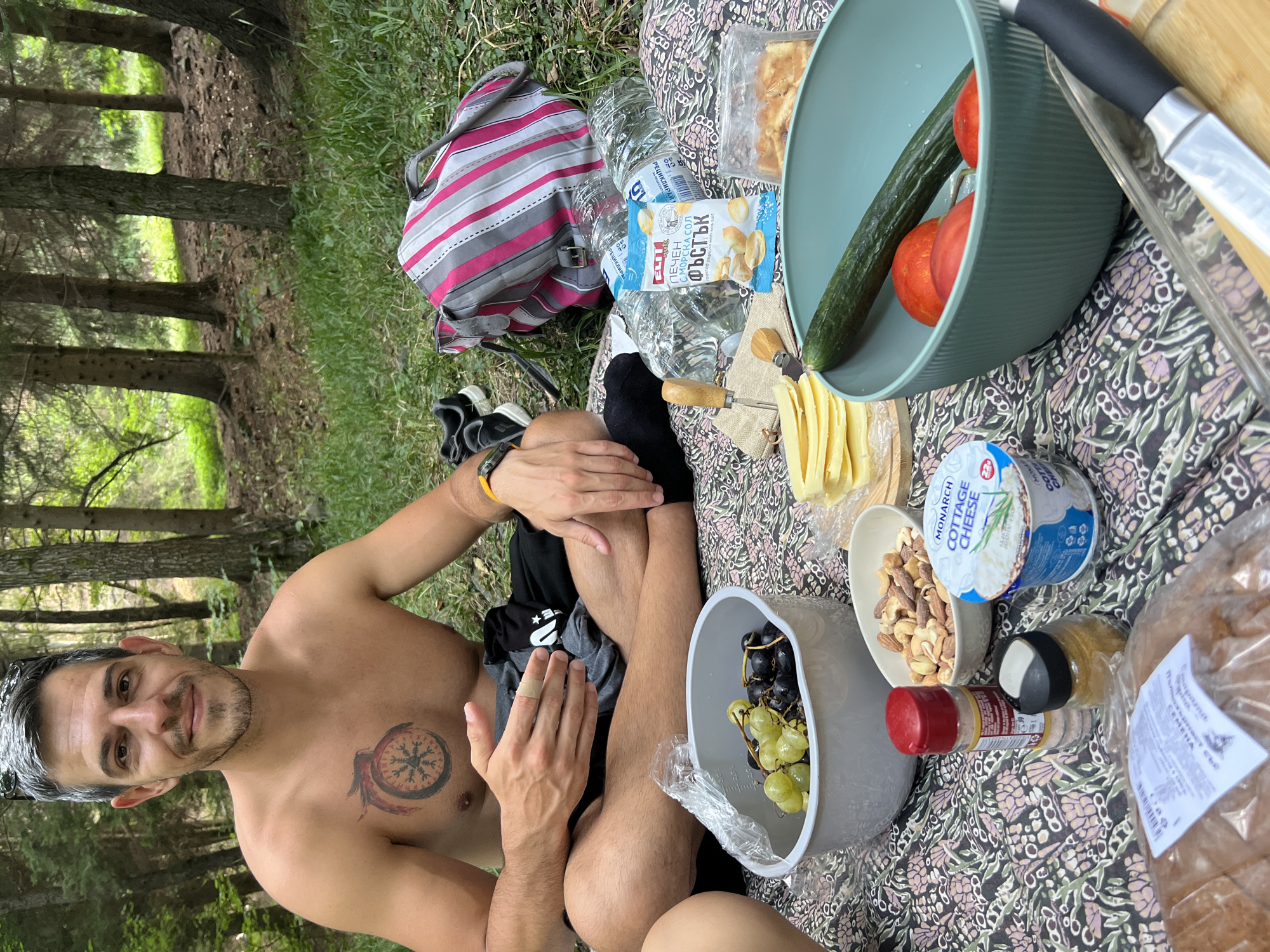 A man with graying hair, a bare chest with a tattoo on his chest, and a beard is sitting cross-legged on a blanket spread on the ground in a forest. He is looking at the camera and smiling. In front of him, there are various food items laid out, including grapes, nuts, cottage cheese, tomatoes, a cucumber, bread, and a packet of crackers. There is also a bottle of water, a striped bag, and a pair of shoes. The setting appears to be a picnic in the woods of Zheleznitsa, Bulgaria.