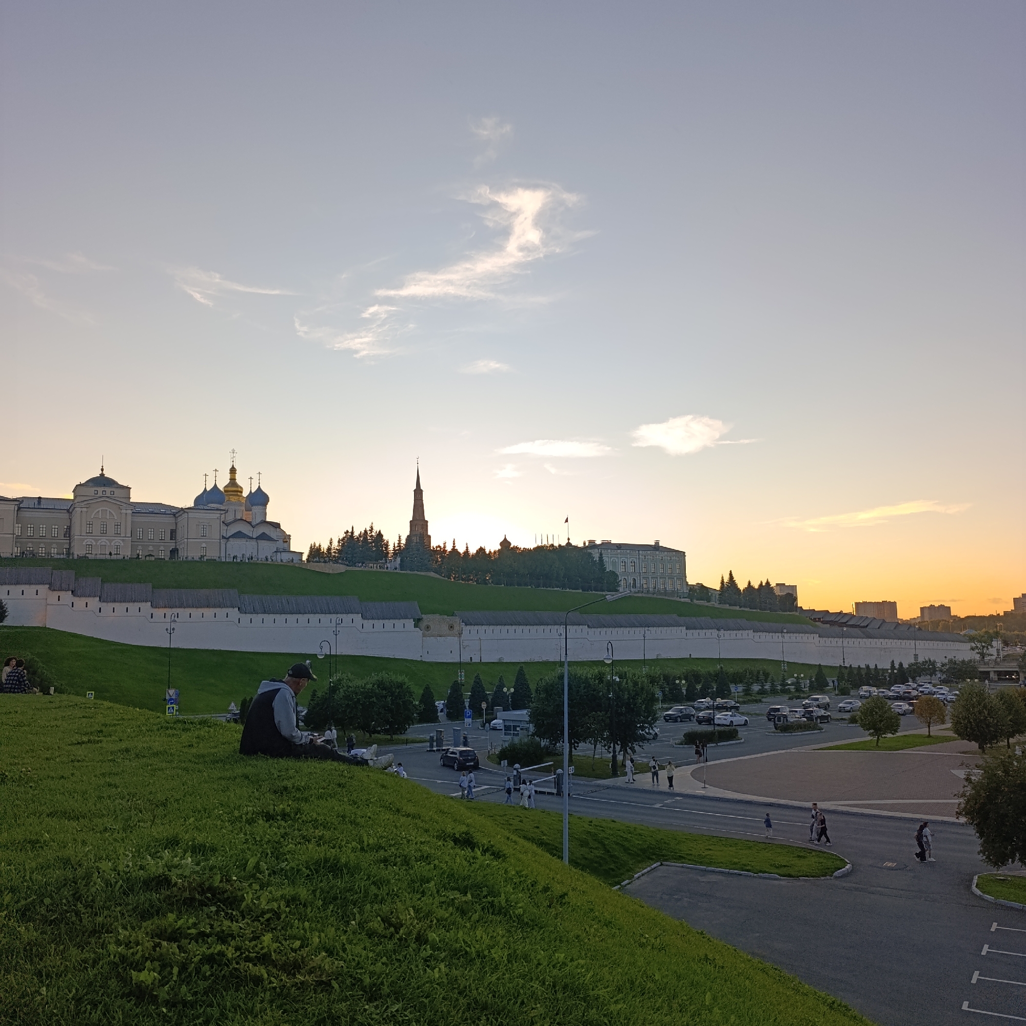 The image depicts a panoramic view of the Kazan Kremlin in Kazan, Russia, at sunset. The Kremlin walls are white and stretch across the hillside, with a distinctive tower, likely the Syuyumbike Tower, visible in the middle distance. Several domed buildings, characteristic of Russian Orthodox churches, are also prominent within the Kremlin complex. The sky is a gradient of soft blues and oranges, indicative of the sunset, with wispy clouds scattered across it.
In the foreground, a lush green grassy slope dominates the lower portion of the frame. A man wearing a baseball cap and a dark vest over a grey hooded sweatshirt is seated on the grass, facing away from the camera and towards the Kremlin, appearing to be resting or observing the scene. Further down the slope, two individuals are seated on the grass, also facing the Kremlin.
Below the Kremlin walls, a road is visible, with cars parked in a lot and driving. Several people are walking along the road and crossing it. Trees and landscaping are interspersed throughout the scene, with a line of coniferous trees bordering the Kremlin. The overall atmosphere is serene and picturesque, capturing a moment of tranquility during the golden hour.