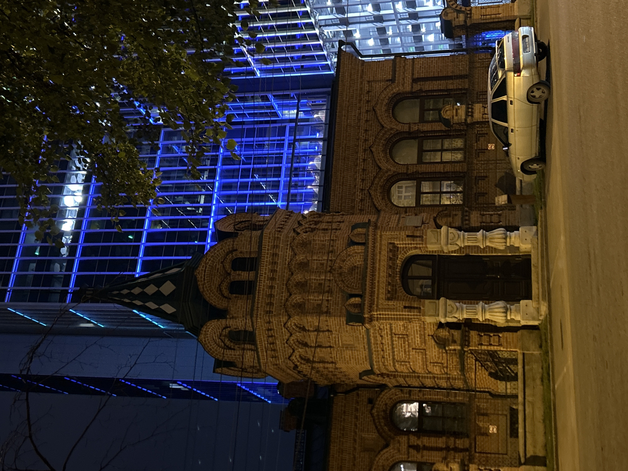 The image depicts a night scene in Yekaterinburg, Russia, featuring an old brick building with distinctive architectural details, including arched windows and a conical turret topped with a diamond-patterned spire. This building is juxtaposed against a modern high-rise skyscraper with a blue, illuminated glass facade. A silver sedan is parked on the asphalt in front of the brick building. A tree with dense foliage is positioned in the foreground, partially obscuring the view of the skyscraper. The street is paved and appears to be a public thoroughfare. The overall atmosphere is urban and nocturnal, highlighting the contrast between historical and contemporary architecture.