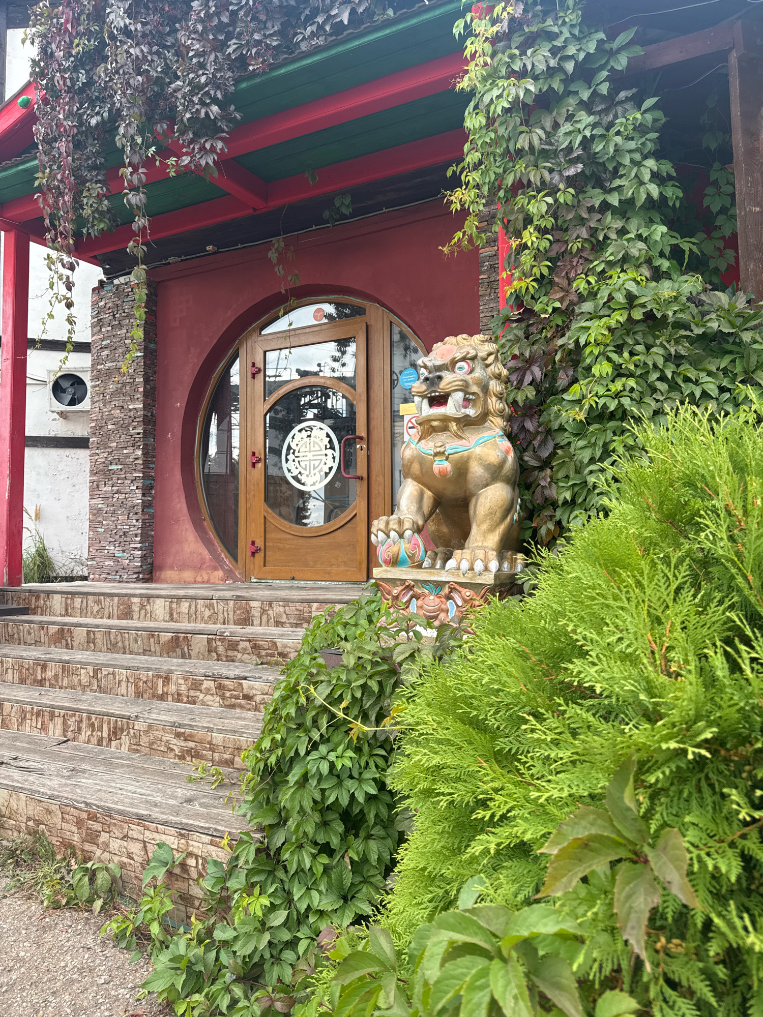 The image shows the entrance to a building with a distinctive red facade and a round wooden door with a white circular design. A stone lion statue, adorned with colorful accents, stands to the right of the entrance. Lush green ivy and other plants climb the walls and overhang the roof. Wooden stairs with stone risers lead up to the entrance. To the left of the door, an air conditioning unit is mounted on a white wall. The scene appears to be an outdoor setting, possibly a restaurant or cultural establishment. This location is the entrance to the restaurant "Kruglaya Ufa" in Ufa, Russia.