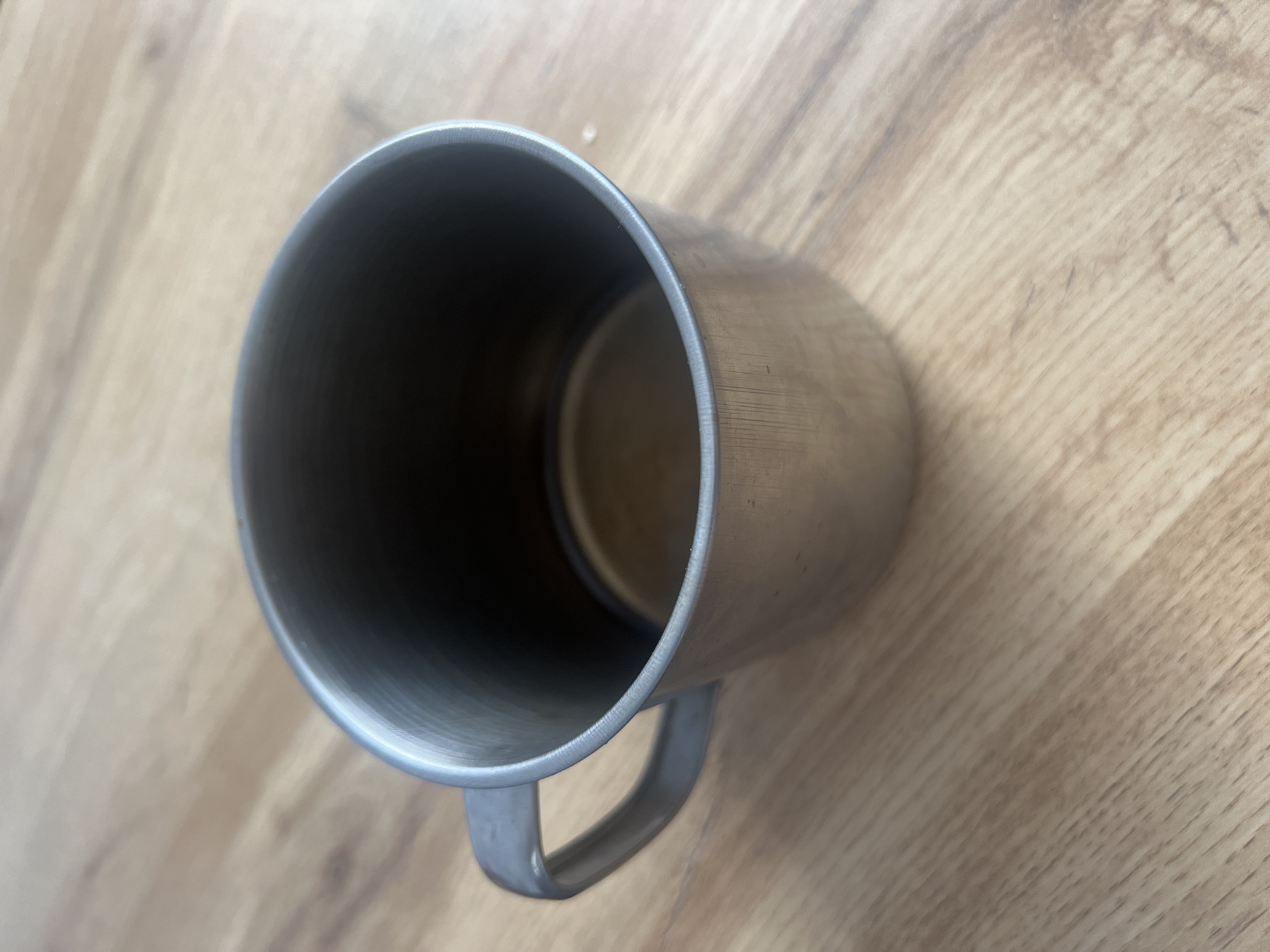 The image shows a metal mug on a wooden surface. The mug is seen from a top-down perspective, tilted slightly to the right. It has a simple, cylindrical shape with a handle attached to one side. The interior of the mug is dark, suggesting it is empty or filled with a dark liquid. The exterior of the mug has a brushed metal finish, and there are some visible scratches or scuffs. The wooden surface appears to be a tabletop or floor with a visible wood grain pattern. The lighting suggests it is daytime, but no specific time of day can be determined. There is no text visible in the image. The scene is simple and straightforward, focusing solely on the object and its immediate surroundings.