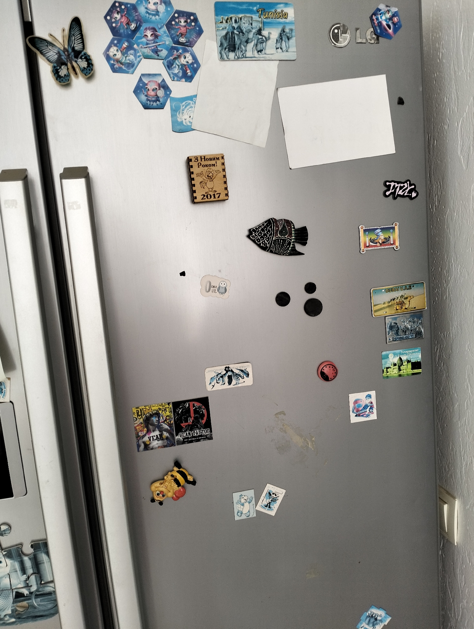 A frontal view of a refrigerator door covered in various magnets and stickers. The door appears to be stainless steel, showing smudges and marks.
On the left side are the handles, partially visible, and a series of hexagonal magnets and a butterfly magnet. There is also a wooden magnet with Cyrillic script and the year "2017."
To the right is a magnet that says "Tunisia," LG logo and several other magnets. There are magnets featuring landscapes, cartoons, and other graphic designs. Also, there are two blank white pieces of paper attached with magnets, a magnet that says "ITZK", and a fish-shaped magnet.
Near the bottom are a few magnets that say "VEAN", "VEAN TATTOO", and a bee-shaped magnet. In the lower right corner, the edge of a wall and a light switch are visible.
The city location in Krykhivtsi, Ukraine is not discernible in the image.