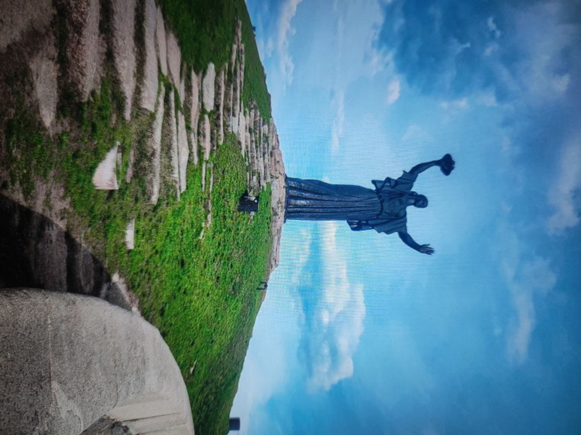 The image shows a statue of a woman with outstretched arms, holding an object in her right hand. The statue is located on a grassy hill with stone steps leading up to it. The background is a clear blue sky with some clouds. The statue appears to be the Motherland Monument in Cherkasy, Ukraine. The monument is a symbol of Ukraine's victory in World War II. The statue is made of bronze and stands on a granite pedestal. The Motherland Monument is one of the most iconic landmarks in Cherkasy.