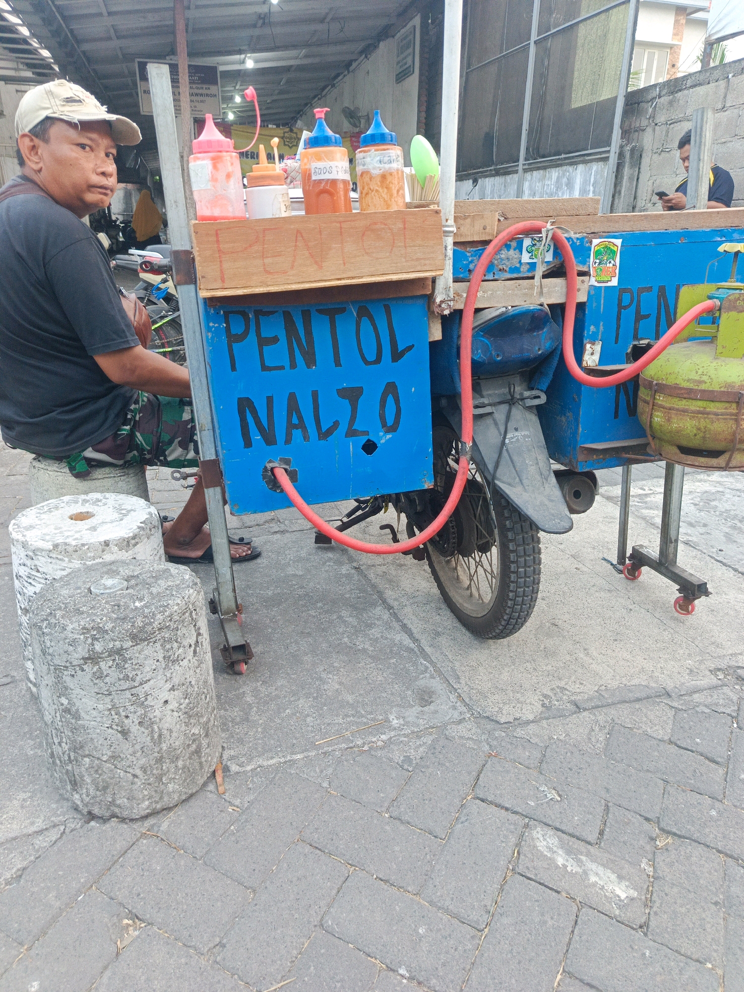 In Pepe, Indonesia, a man in a beige cap and dark t-shirt sits on a large cylindrical concrete object to the left of the frame. He is looking towards the viewer. He is wearing camouflage patterned shorts. In front of him is a food cart, primarily painted blue, mounted on a motorcycle chassis. The cart has "PENTOL NALZO" painted in large black letters on its side. Several bottles of condiments are visible on the top of the cart. A red hose runs from the cart to a green LPG cylinder on the right side of the frame. The cart has a wooden front panel and what appears to be a food preparation area. The scene takes place on a paved street with some debris. In the background, another man is seated and looking at a phone, with buildings and fencing visible.