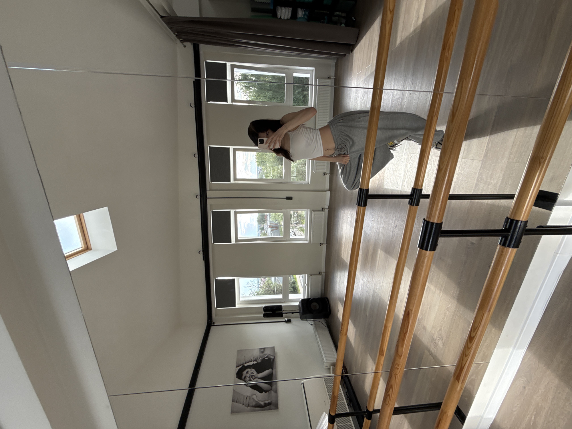 A full-length mirror reflects a young woman taking a selfie in a dance studio. The woman is wearing a white crop top and grey sweatpants. She is posing with one leg bent and her arm extended, holding a phone. The studio has wooden floors, a barre for dancing, and large windows. A skylight is visible in the ceiling. Artwork hangs on the wall, depicting ballet dancers. Speakers and radiators are also present in the room. In the reflection, the woman is positioned in the center of the room. The scene suggests a dance or fitness environment.