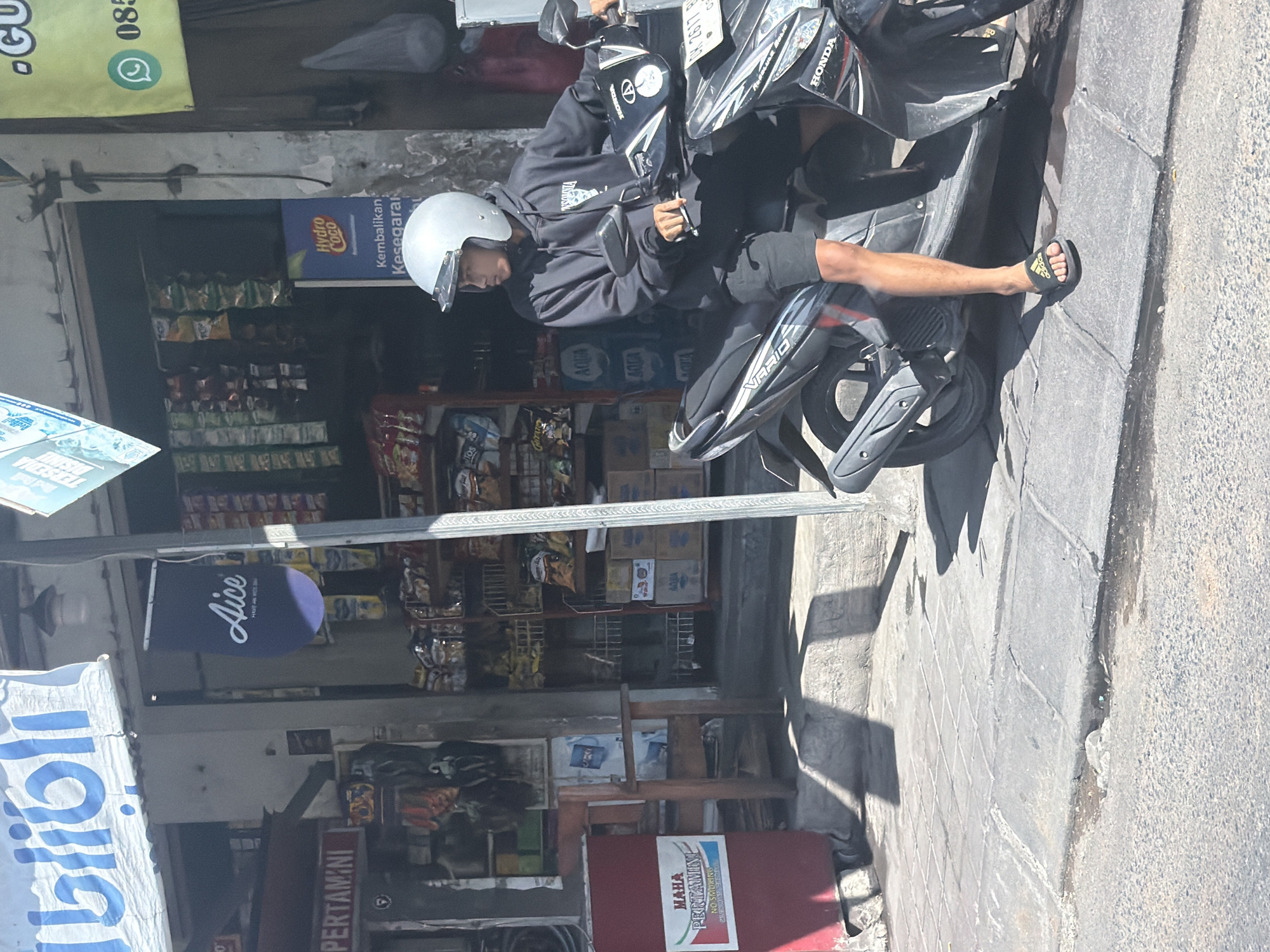 The image, taken in North Kuta, Indonesia, depicts a bright daylight scene featuring a person on a parked scooter beside a street-side shop. A person wearing a dark hoodie, shorts, and a white helmet is seated on a black Honda Vario scooter. Their right foot, clad in an Adidas slide, rests on the concrete sidewalk. The scooter's license plate, "DK 2617 RAO", is visible.
Behind the scooter is a small, open-front convenience store or warung, its shelves stocked with various products including snacks (like "CHITATO" and "TOS") and bottled water ("AQUA"). Multiple signs and banners adorn the shop's facade: one for "Hydro Coco" with the slogan "Kembalikan Kesegaran" (Restore Freshness), an "Aice" sign, and a prominent "MAHA PERTAMINI NO SMOKING" sign, suggesting a small fuel vendor. Other visible text includes "Blibli" on a hanging banner and "GU 085" on another, likely a contact number. The scene is illuminated by bright sunlight, casting sharp shadows on the concrete sidewalk and asphalt road, indicating a clear day. The overall ambiance is that of a casual, everyday street scene in a warm climate.