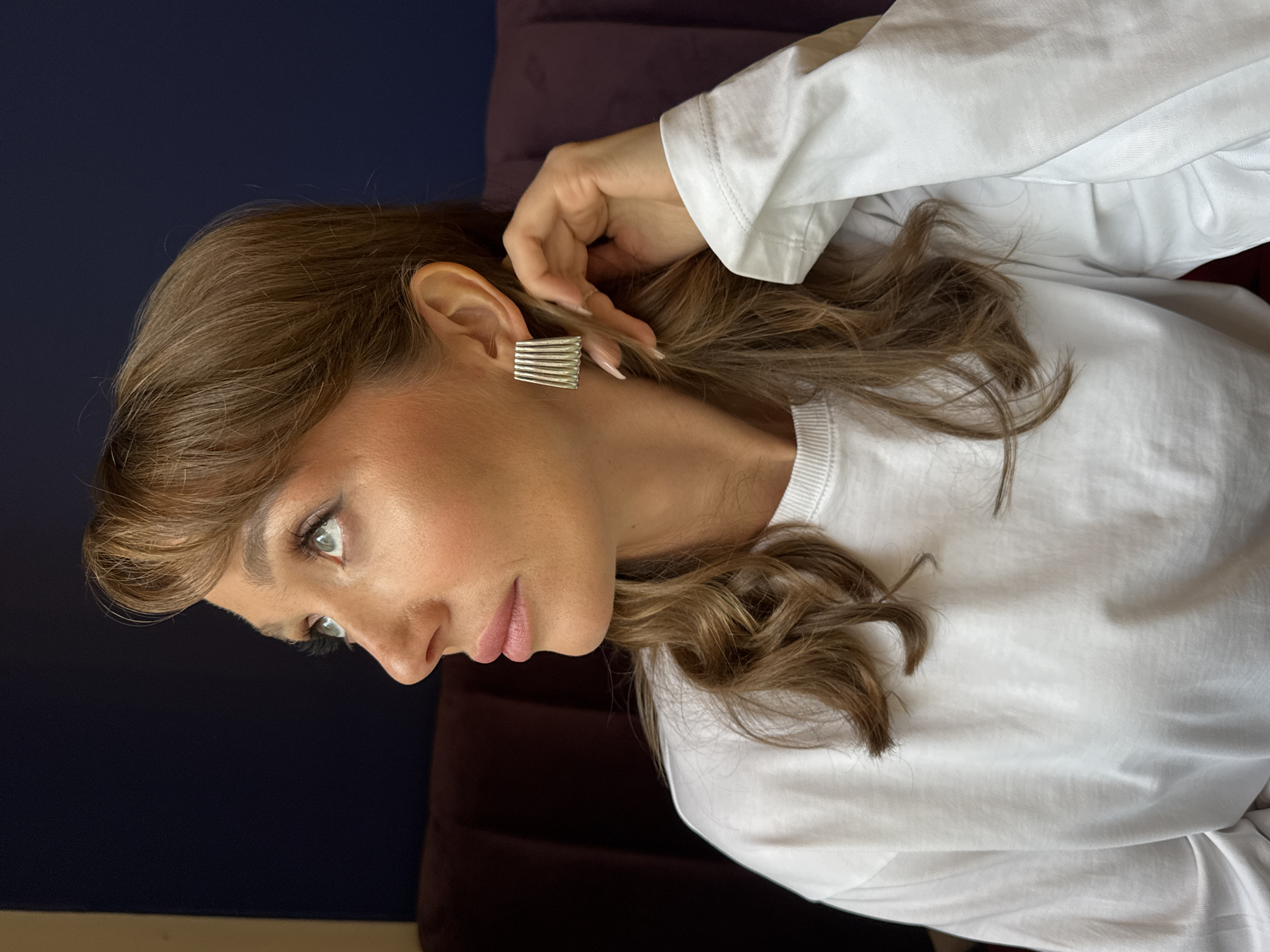 A woman with light brown hair and green eyes is seen in a close-up, medium shot, facing left. She is wearing a white, long-sleeved shirt and a silver, rectangular earring. Her right hand is raised towards her ear. The background consists of a dark blue and a dark purple surface. There is no indication of this being in Saint Petersburg, Russia.