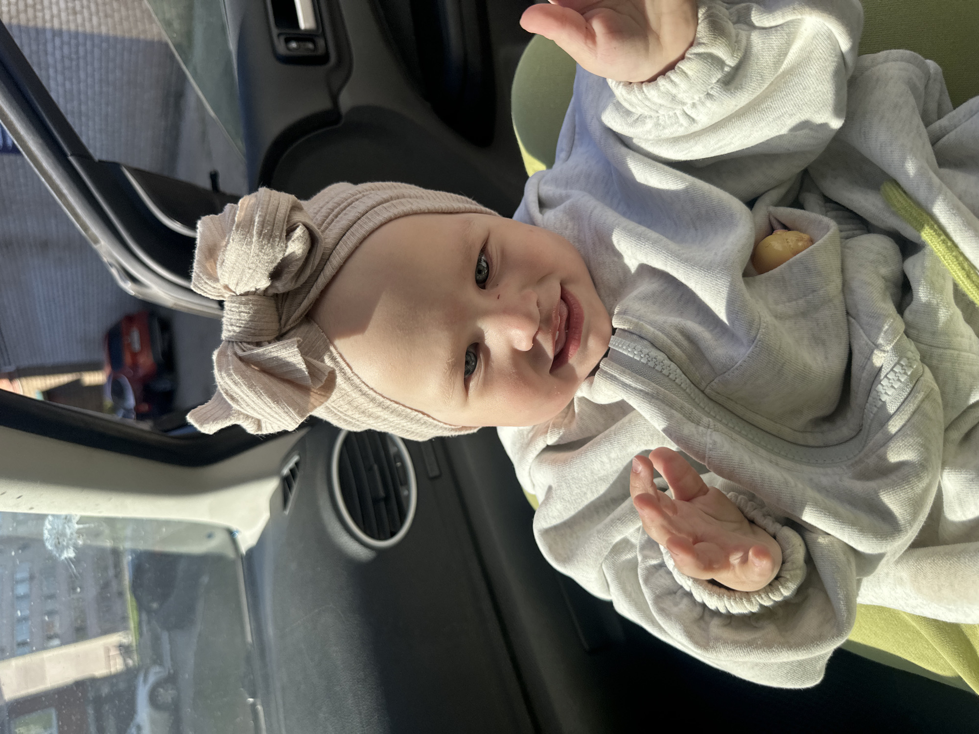 A baby with a beige headband and a gray sweater is sitting in a car seat in the front passenger seat of a car. The baby is facing the camera with its mouth slightly open and hands raised. The car's dashboard and part of the windshield are visible. Outside the car, a building and trees are visible. The location appears to be a city street. There is no indication that this photo was taken in Chaikovsky, Russia.