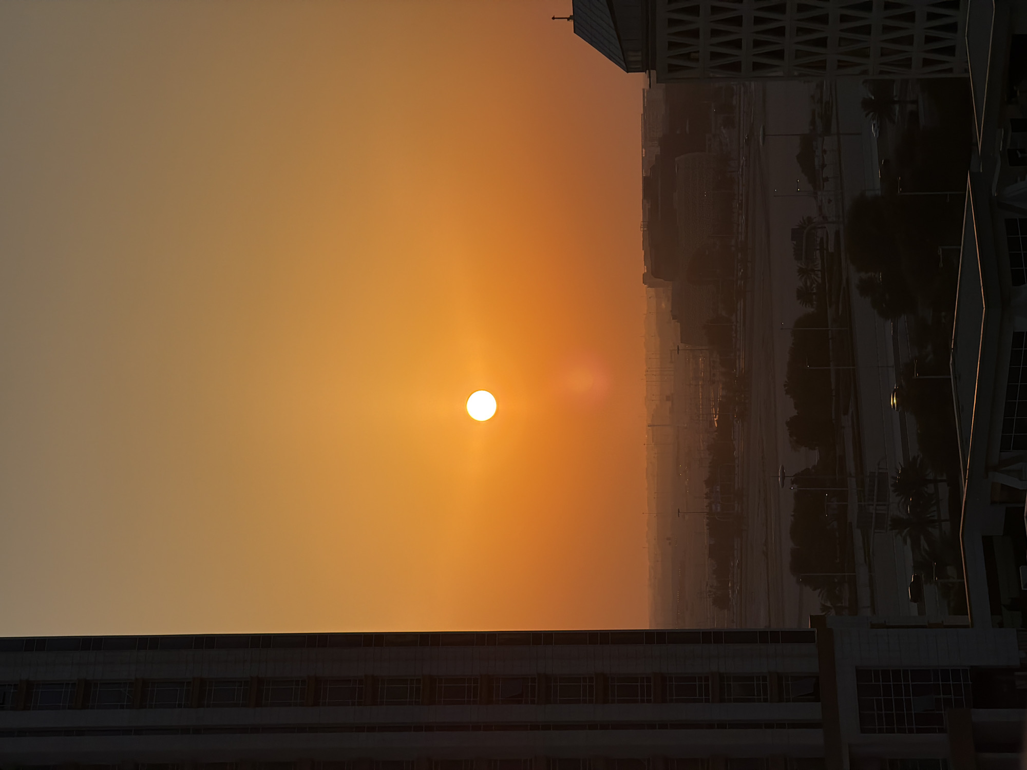 The image depicts a sunset over a city skyline. The sun, a bright white disc, is low in the sky, casting a warm orange glow across the atmosphere. Tall, modern buildings frame the scene, their geometric shapes silhouetted against the vibrant sky. One building in the foreground, on the left, is dark and features rows of windows. Another building, on the right, has a more complex facade with a latticed pattern. In the distance, more structures and palm trees are visible, suggesting an urban environment. Streetlights and pathways are faintly discernible, hinting at the city's infrastructure. The overall mood is serene and tranquil, characteristic of a sunset. The time of day is clearly evening, as indicated by the sun's position and the ambient light. There is no visible text in the image.