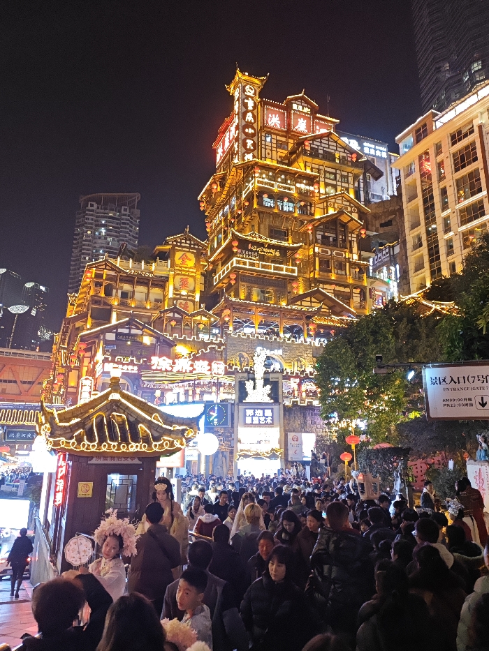 The image depicts a bustling night scene at Hongyadong scenic area, located in Yuzhong District, Chongqing, China. The focal point is a multi-tiered, traditional Chinese stilt-house complex, intensely illuminated with golden lights that highlight its intricate wooden architecture, multiple balconies, and pagoda-style roofs. Prominent vertical neon signage displaying Chinese characters glows brightly on the main structure. To the right and behind, modern high-rise buildings with illuminated windows contrast with the traditional design.
In the foreground and midground, a dense crowd of people, predominantly East Asian adults and children, fills the street and plaza areas. They are walking, standing, and observing the illuminated environment. Two individuals in elaborate traditional Chinese costumes are visible in the foreground: one person wears a large white, fan-like headpiece and light-colored attire, smiling towards the viewer, while another in golden robes and an ornate headdress stands elevated, possibly performing or posing for photos.
The immediate environment includes smaller traditional-style booths and structures adorned with lights. Green trees and foliage on the right are also strung with decorative lights. A white sign with black text on the right indicates "景区入口 (7号门)" (Scenic Area Entrance (Gate 7)) and operating hours: "AM 09:00 开园 (Open) / PM 23:00 闭园 (Closed)". Red lanterns hang from branches and structures. The overall scene conveys a lively, vibrant atmosphere in a popular cultural and commercial district.