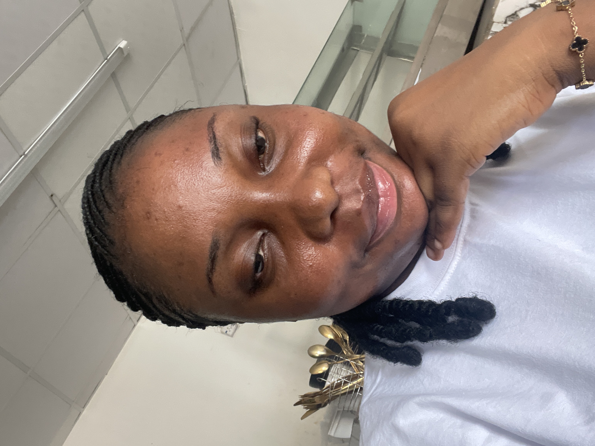 A close-up portrait of a woman with dark skin and braided hair. She is wearing a white t-shirt and a gold bracelet. Her hand is positioned near her face. In the background, there is a wall with a grid pattern and a metal frame. The presence of the spoons and forks would suggest the image was taken in a restaurant or a cafeteria. There is no evidence that can verify if this image was taken in the city of Jos, Nigeria.