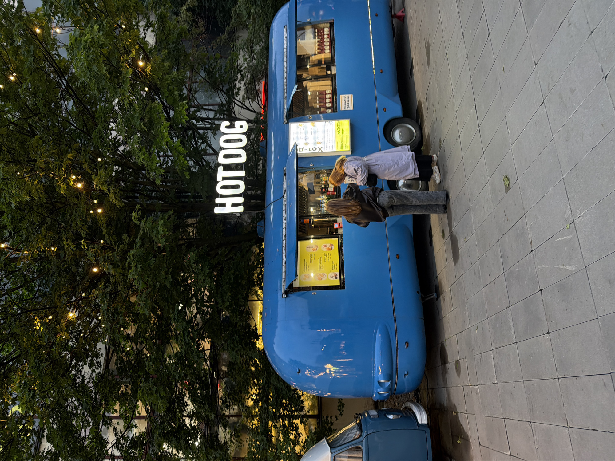 The image shows a blue food truck named "HOT DOG" parked on a paved area. Two young women are standing at the service window of the food truck, appearing to order food. The food truck has a retro design, and there is another blue vehicle, possibly a van, parked to its left. String lights are visible in the trees behind the food truck, suggesting it is evening or nighttime. The background includes a building with reflective windows. The overall setting appears to be an outdoor urban environment.