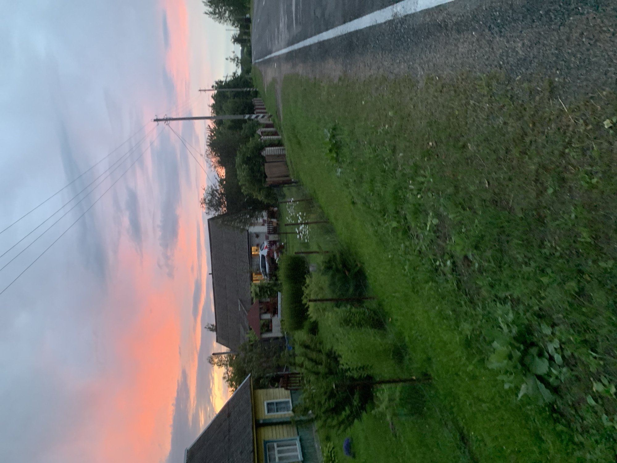 The image depicts a rural scene in Belarus at dusk. The sky is filled with soft pink and orange hues, indicating the sunset. In the foreground, a grassy area borders a paved road with a white line. Beyond the grass, there are several houses with dark roofs and yellow and grey siding. Some houses have windows with white trim. A fence runs along the properties. In one of the yards, a white car is parked, and the lights are on. The overall setting suggests a quiet village or countryside. There is no visible text. The scene evokes a peaceful and serene atmosphere as day transitions into night.