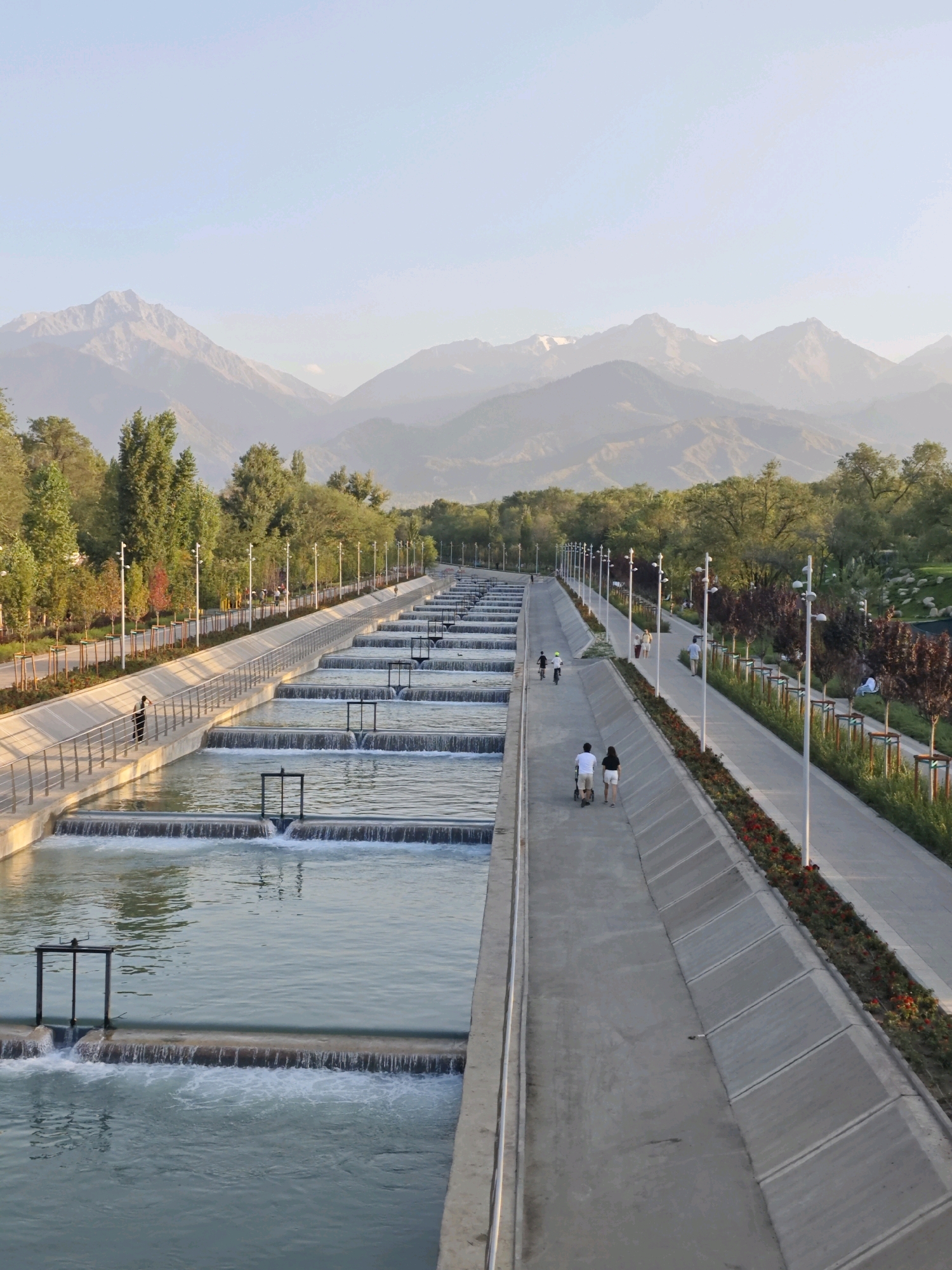 The image shows a cascading water feature alongside a pedestrian walkway with a mountain range in the background. The water feature consists of a series of concrete channels with small waterfalls, creating a terraced effect. The walkway is lined with trees, lampposts, and flower beds. Several people are visible on the walkway, some walking and others cycling. In the foreground, a person stands near the railing of the water feature. The scene appears to be a park or recreational area within a city, likely Almaty, Kazakhstan, given the prominent mountain backdrop. The sky is clear and pale blue, suggesting daytime.