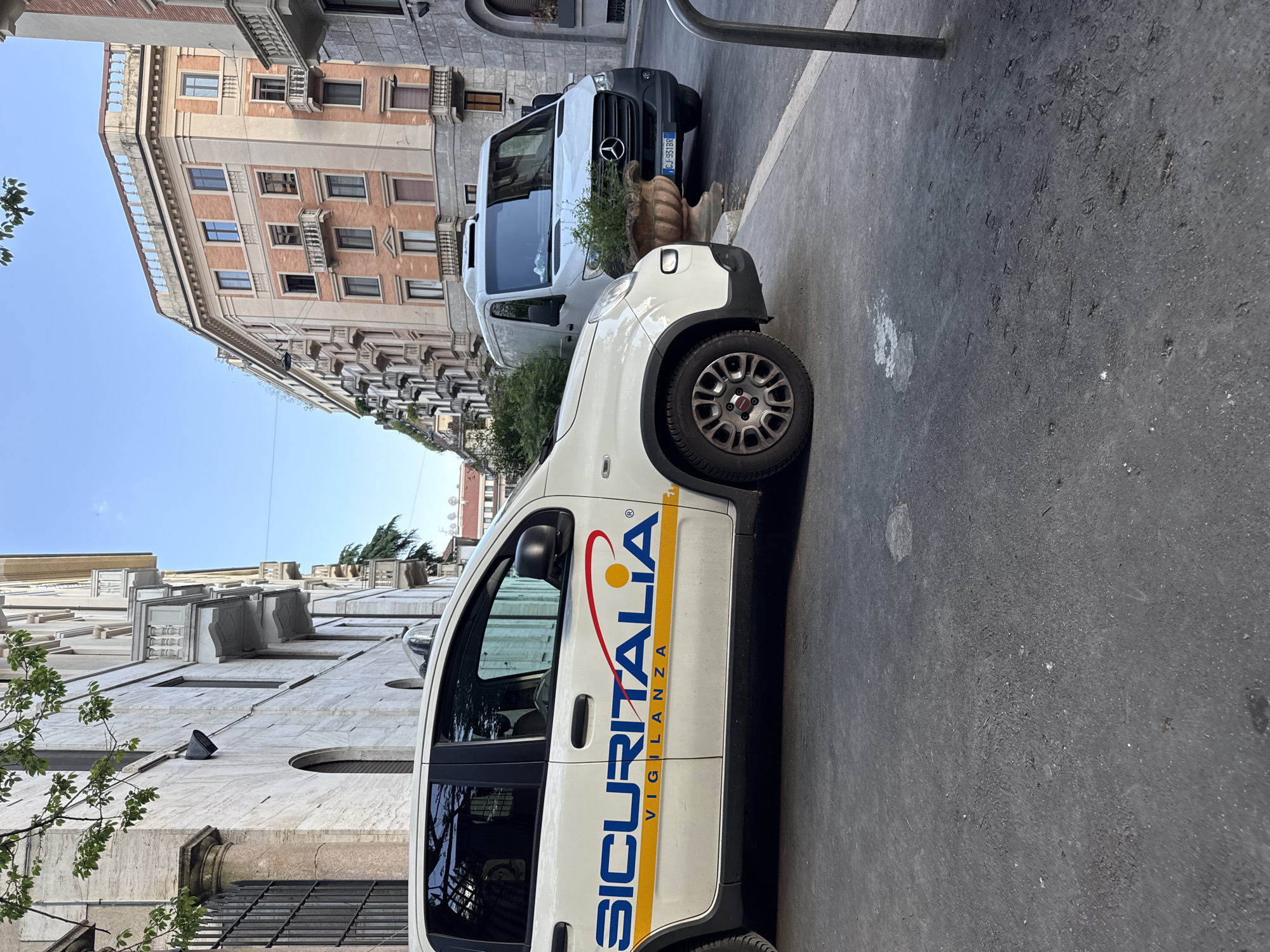 The image displays a street scene in Milan, Italy, captured on a clear, sunny day. In the foreground, a white Fiat Panda utility vehicle is parked, featuring the prominent blue and red logo of "SICURITALIA VIGILANZA" (security surveillance) on its side, along with a registered trademark symbol.
Behind the white security car, a white Mercedes-Benz Sprinter van is also parked, with its front visible. Its Italian license plate, "CJ 991BR," is clearly readable. Between the two vehicles, a decorative stone planter holds some green shrubbery. The vehicles are situated on an asphalt street next to a sidewalk.
On the left side of the street, a multi-story building with classic European architecture dominates the background. It features a terracotta-colored facade with numerous windows and balconies, indicative of residential or commercial properties. Further down the street and to the right of the security car, another impressive building with light-colored, possibly stone or marble, cladding and decorative architectural elements is visible. Patches of green foliage from trees or bushes are present in the scene, adding natural elements to the urban environment. The bright blue sky suggests midday sun.