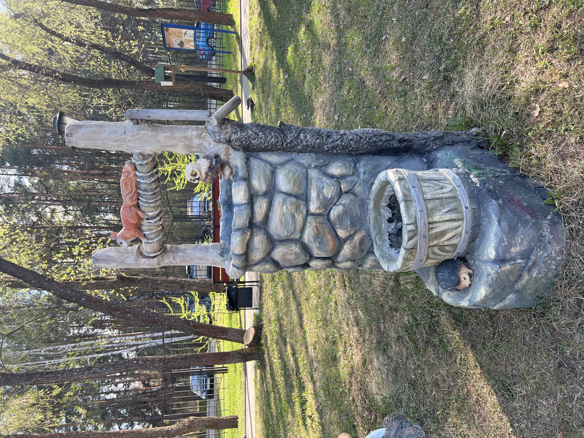 The image shows a decorative monument in a park in Chaikovsky, Russia. The monument is made of artificial materials and painted to resemble a stone well and a tree stump. A faux squirrel, owl, and hedgehog are incorporated into the design. The surrounding environment includes trees, grass, and a metal fence. A road with parked cars can be seen in the background.