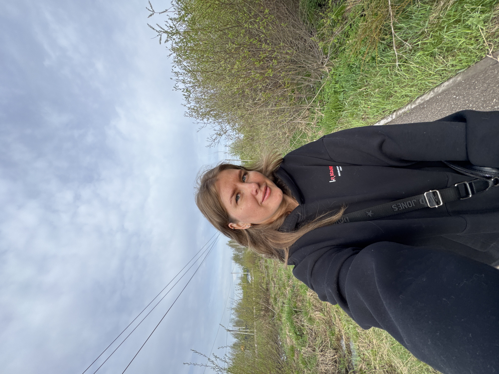 A woman with blonde hair is standing on a path or roadside in Ферма, Russia, taking a selfie. She is wearing a black hoodie with a logo in red and black, and a shoulder bag with the name "D JONES" printed on the strap. The background consists of a cloudy sky, foliage including shrubs or bushes, and power lines. The immediate surroundings appear to be a rural or suburban area.