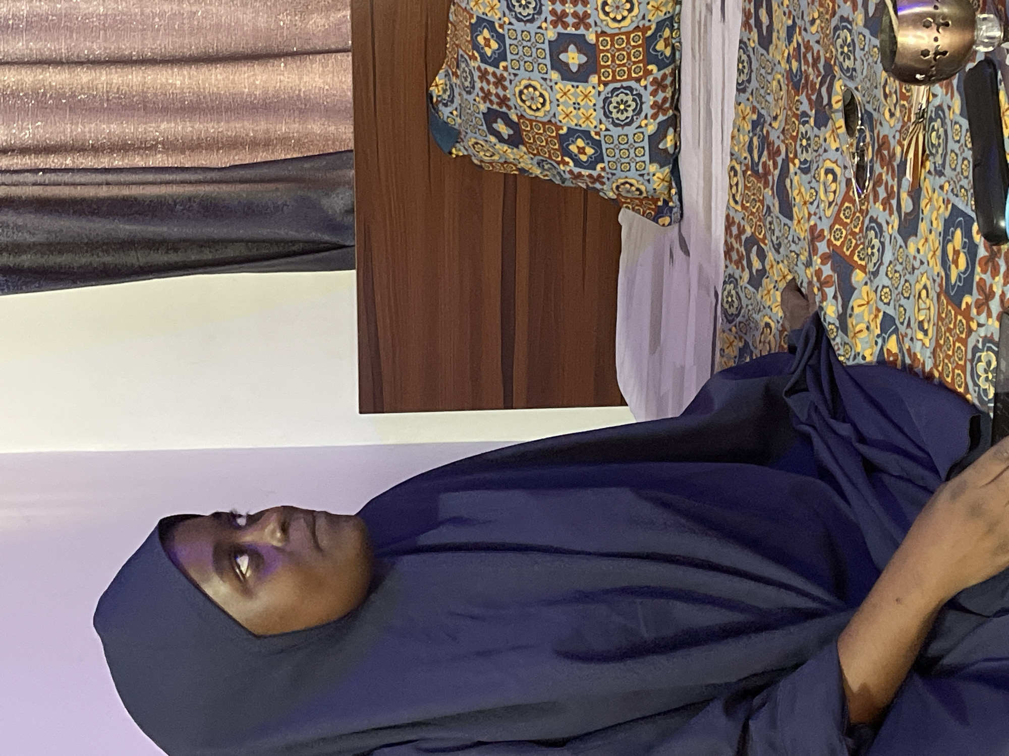 A person wearing a dark blue hijab lies supine. The background includes a wooden cabinet, patterned fabric, and a silver-colored object. No location information is verifiable.