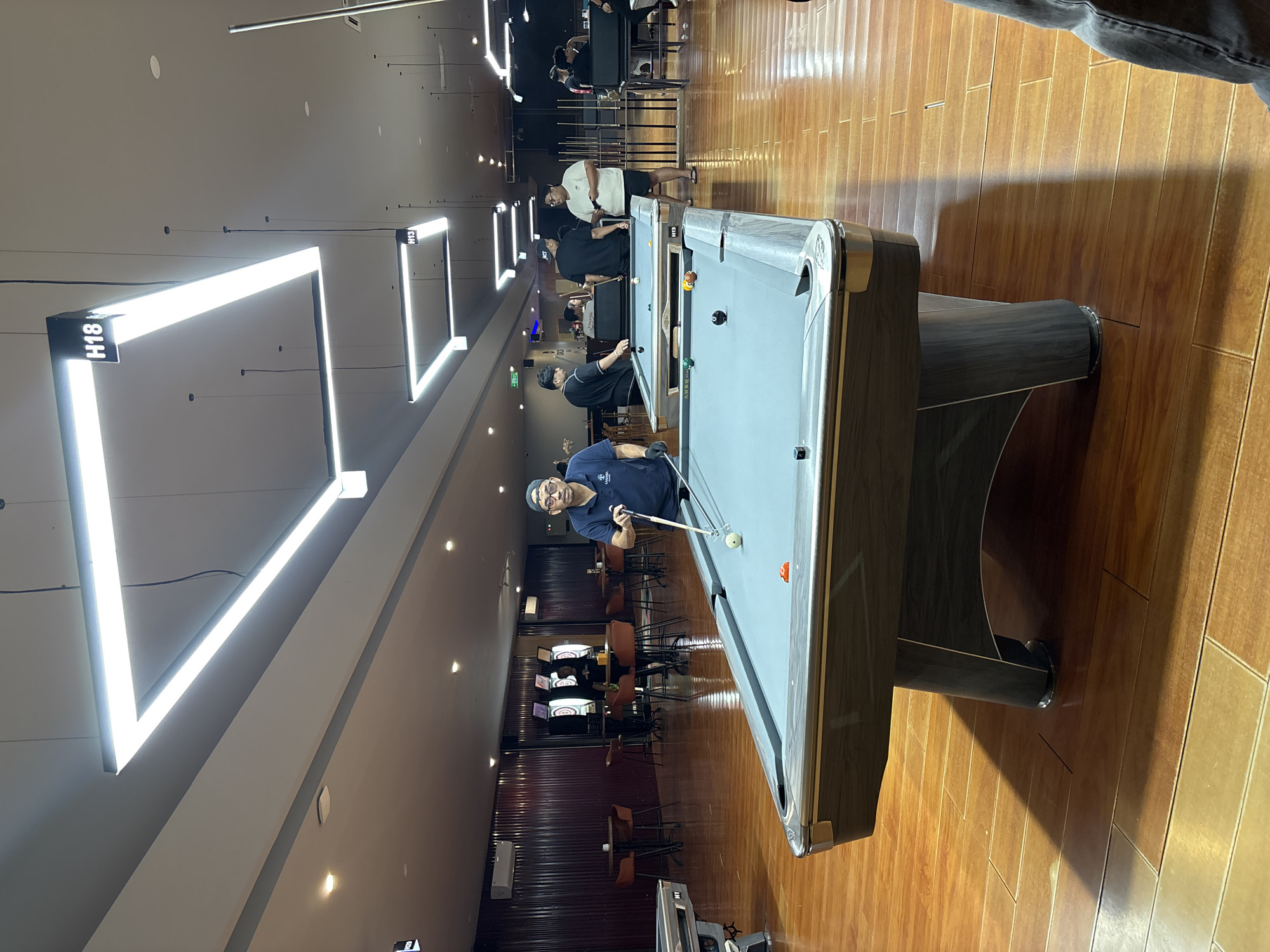 This image depicts the interior of a modern billiard hall, possibly located in Kuta Selatan, Indonesia. The scene is illuminated by prominent rectangular fluorescent light fixtures suspended from the ceiling. In the foreground, a grey pool table with a patterned wooden frame is centrally positioned. One pool ball, orange in color, rests on the table's surface. A man wearing a dark blue polo shirt, a blue cap, and black gloves is standing to the left of this pool table, holding a pool cue. He is in the process of playing.
Further into the background, other pool tables are visible, some with people engaged in playing. Several individuals are casually dressed, some standing and observing, others actively participating in games. The environment features dark wooden flooring and dark-colored walls, with some sections having a ribbed texture. In the far background, dartboards are mounted on the wall, suggesting a multi-recreation facility. The overall atmosphere suggests a social gathering or a recreational establishment.