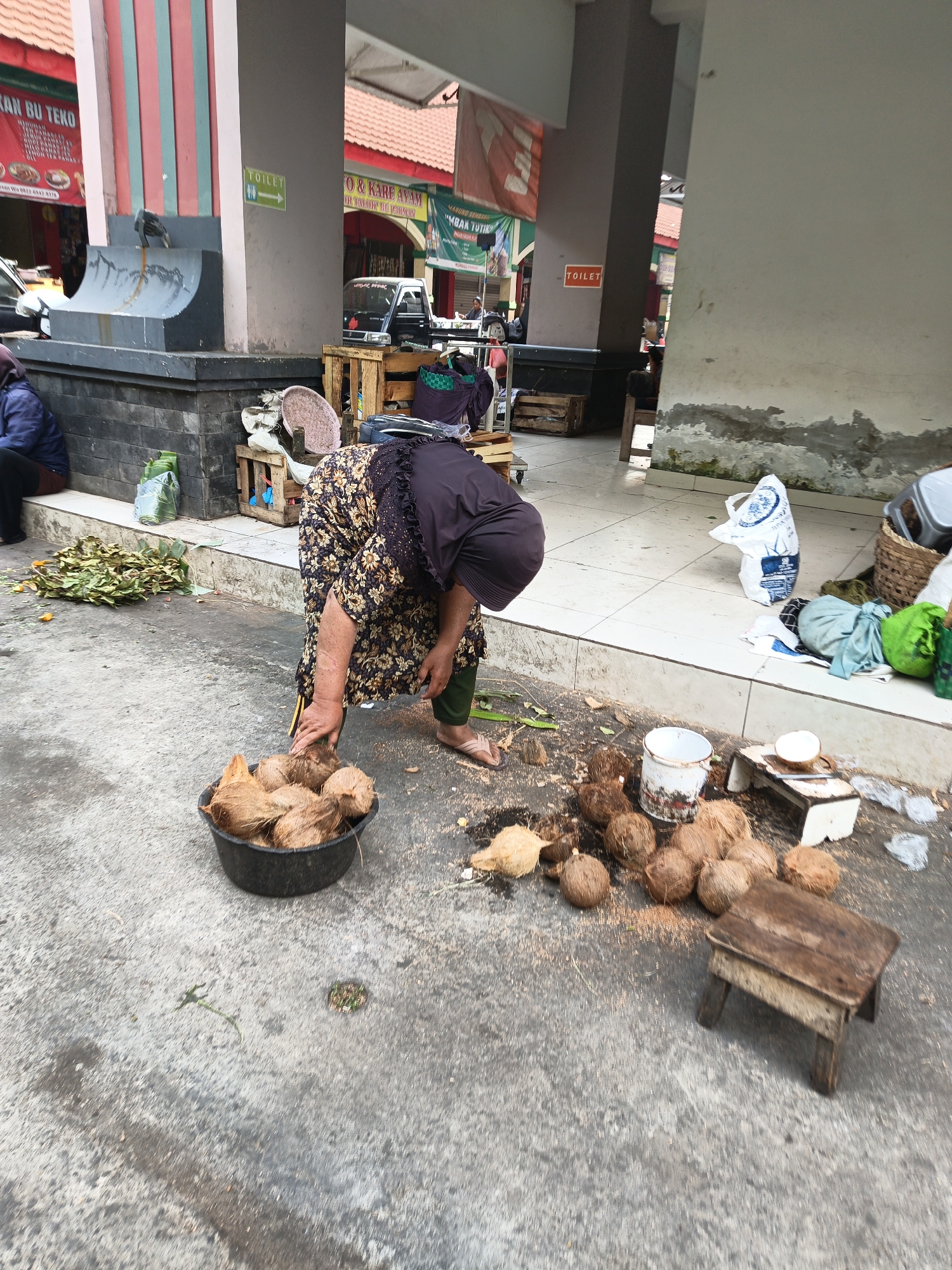 The image depicts an outdoor market scene in Klaten, Indonesia. A woman, dressed in a dark floral-patterned dress and green trousers, is bending over a black bucket filled with coconuts. She is interacting with the coconuts, possibly sorting or preparing them. Scattered around her on the concrete ground are more coconuts, some of which appear to have been opened or are in the process of being opened. There's also a small wooden stool and a white bucket visible. In the background, there are market stalls, signage (including a "TOILET" sign), and a small vehicle. A few other people are partially visible in the background. The environment suggests a typical bustling marketplace with goods laid out for sale.