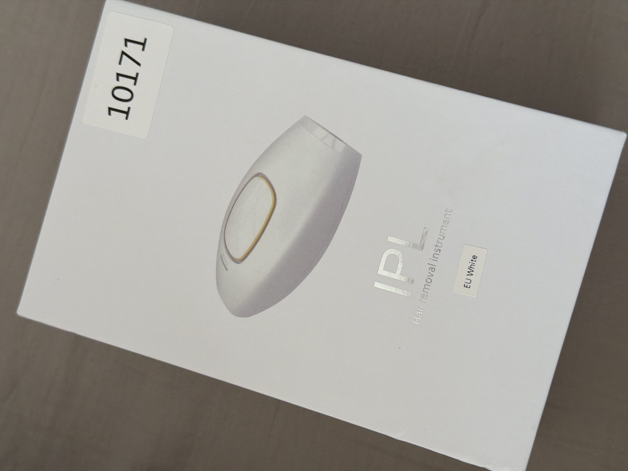 The image shows the top-down view of a white box for an IPL hair removal instrument. The box is on a brown, textured surface. A white sticker with the black numbers "10171" is affixed to the top right corner of the box. The front of the box features a silver, holographic logo for "IPL" and the text "Hair removal instrument" below it, also in silver. Below that, there is a smaller white sticker with the text "EU White". A white IPL hair removal device is pictured in the center of the box. The lighting is even and there are no shadows cast by the box or the device. The image appears to be taken indoors in a studio setting, with no discernible background or environmental context.