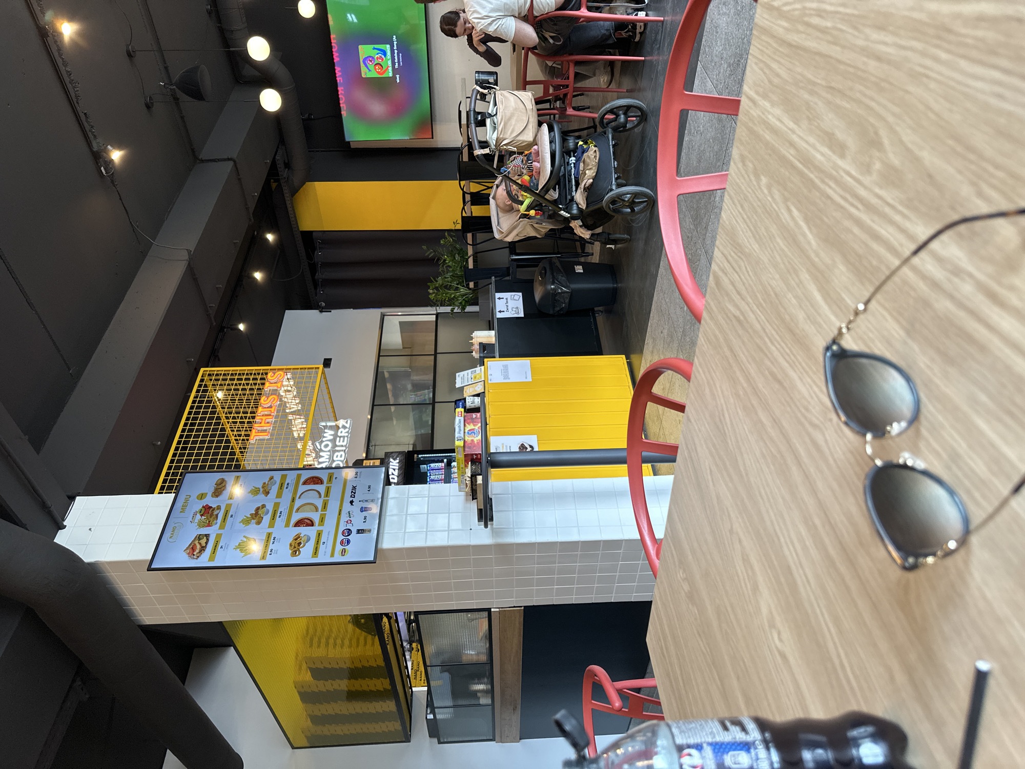 This image depicts the interior of a restaurant, likely located in Wrocław, Poland, as evidenced by Polish text on the menu board. The scene includes a digital menu board displaying food items with prices, a yellow counter, and red chairs. A woman and a baby in a stroller are visible in the background. Sunglasses and a partially visible bottle of dark liquid, possibly soda, are on a wooden table in the foreground. The restaurant has a modern interior design with exposed ductwork and black and yellow accents. A TV screen in the background displays a cartoon.