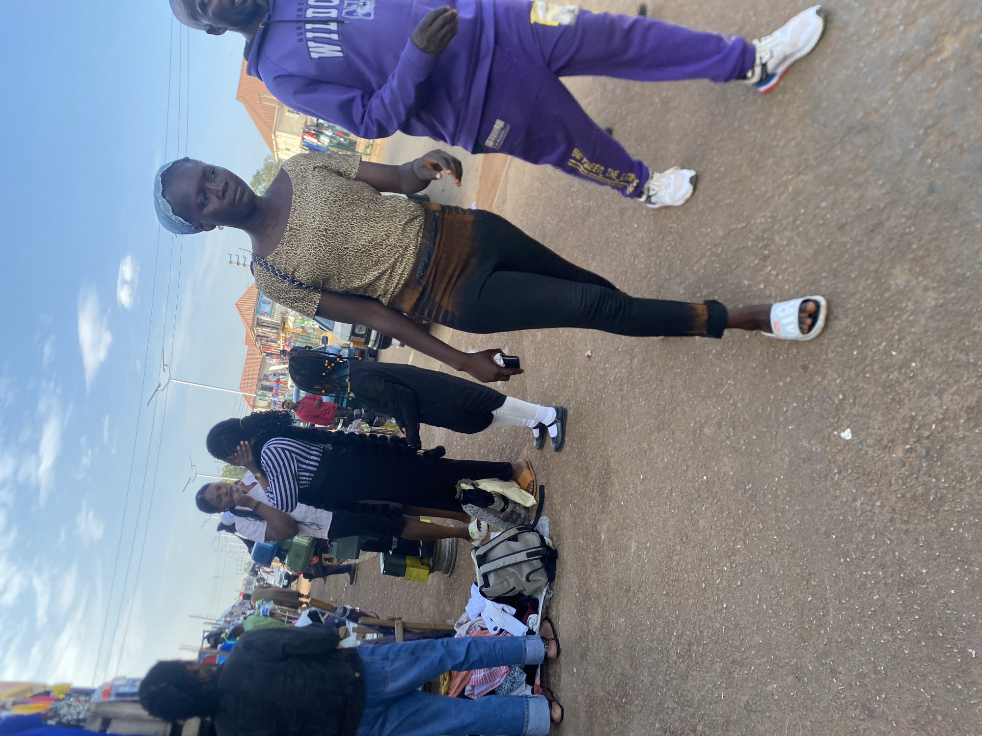 The image captures an outdoor street scene in Bukuru, Nigeria. A woman wearing a leopard-print top and black pants stands in the foreground, with another person wearing a purple outfit partially visible above her. Along the side of the street, a row of people are positioned behind makeshift stalls displaying various items, likely for sale. Some of these people appear to be interacting with one another. The street itself appears to be paved. The sky is visible in the background, showing a clear day.