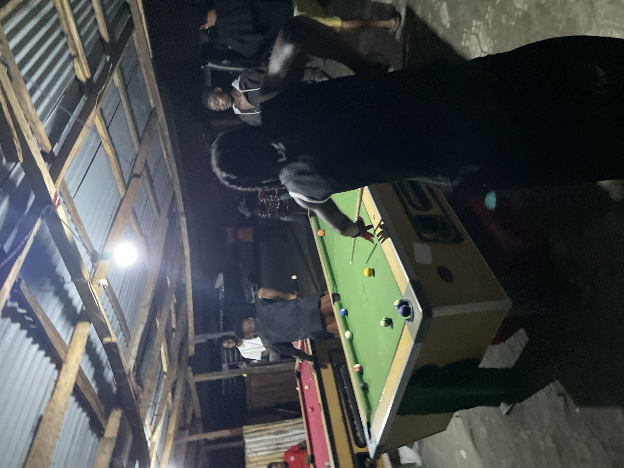 The image depicts an indoor scene at night, likely in Ado, Nigeria, where several men are gathered around a pool table. The setting appears to be a rustic or informally constructed space, with walls and possibly the roof made of corrugated metal sheets, supported by exposed wooden beams and rafters.
In the foreground, a man wearing a dark shirt is actively playing pool on a green-felt table, holding a cue and preparing to strike one of the various colored balls. Another man in a dark shirt and light shorts stands to his left, observing. Several other individuals, all men, are visible in the background, some standing and others possibly seated, engaged in conversation or watching the game. A second pool table, with a red felt surface, is partially visible behind the main one.
The environment is dimly lit by a single, bright, unshielded light bulb suspended from the wooden ceiling structure, creating a focused illumination on the pool table while leaving the surrounding areas in shadow. The floor appears to be made of concrete or rough ground. The overall atmosphere suggests a casual, social gathering centered around the game of billiards. No distinct text is legible within the frame.