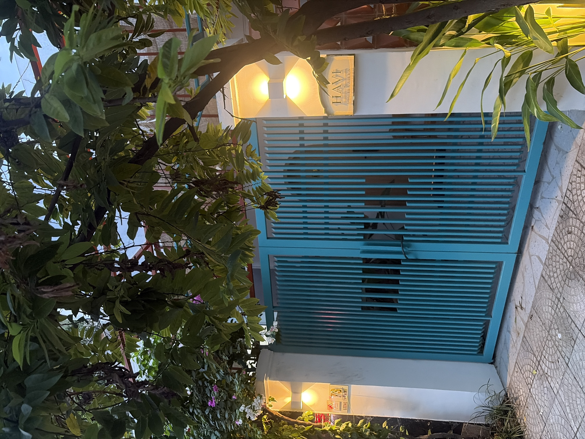 The image shows a turquoise metal gate with vertical slats, flanked by white pillars. Two modern wall sconces with warm yellow light are mounted on the left pillar, and one on the right. A rectangular sign with the text "HAVI" and "SONTRA VILLA" is attached to the right pillar. The gate is partially obscured by lush green foliage from trees and plants. The ground in front of the gate is paved with decorative tiles. In the upper left corner of the left pillar, there is a smaller sign with the number "73" and some Vietnamese text, possibly an address. The scene appears to be in a residential or commercial area in Đà Nẵng, Vietnam, given the architectural style and vegetation. The lighting suggests it is either dusk or dawn. There are no people or animals visible in the image.