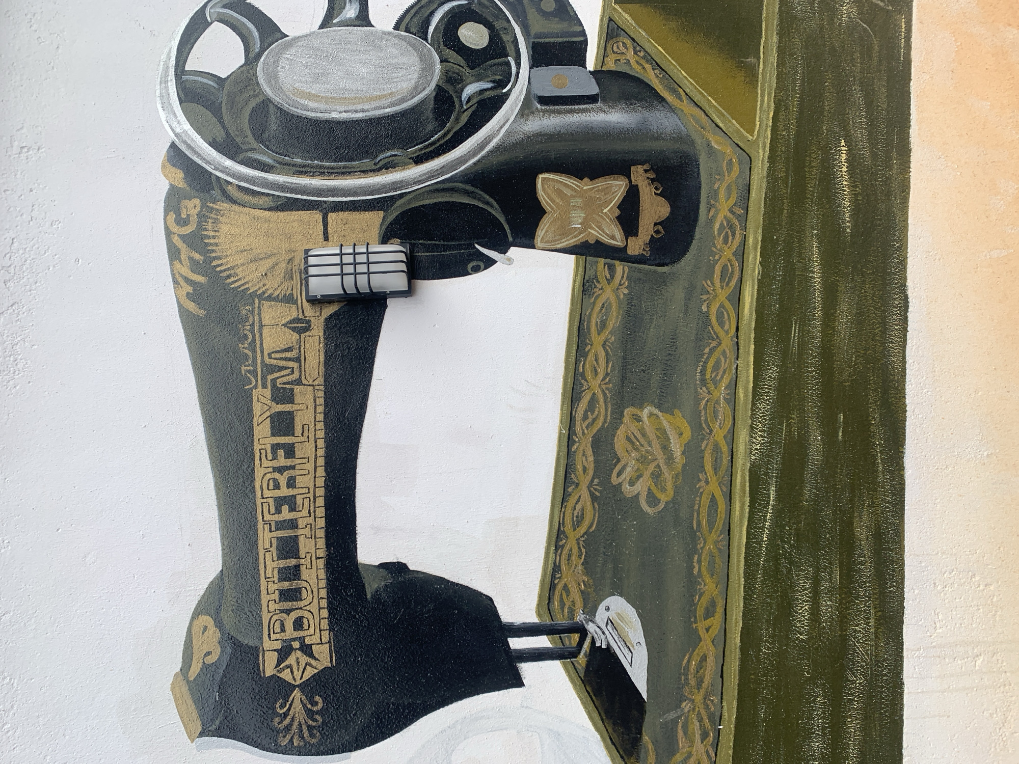 The image is a close-up mural painting of a vintage sewing machine. The sewing machine is depicted in black and gold, with intricate details. The brand name "BUTTERFLY" is prominently displayed in gold lettering on the machine's arm. A metallic thread spool and a handwheel are visible on the right side. The machine rests on a dark green base with ornate gold patterns. The background is a textured white wall. The mural appears to be located on an exterior wall, as suggested by the texture and the hint of a light fixture attached to the machine in the painting. The description of Gura Top, Nigeria is not present in the image itself.