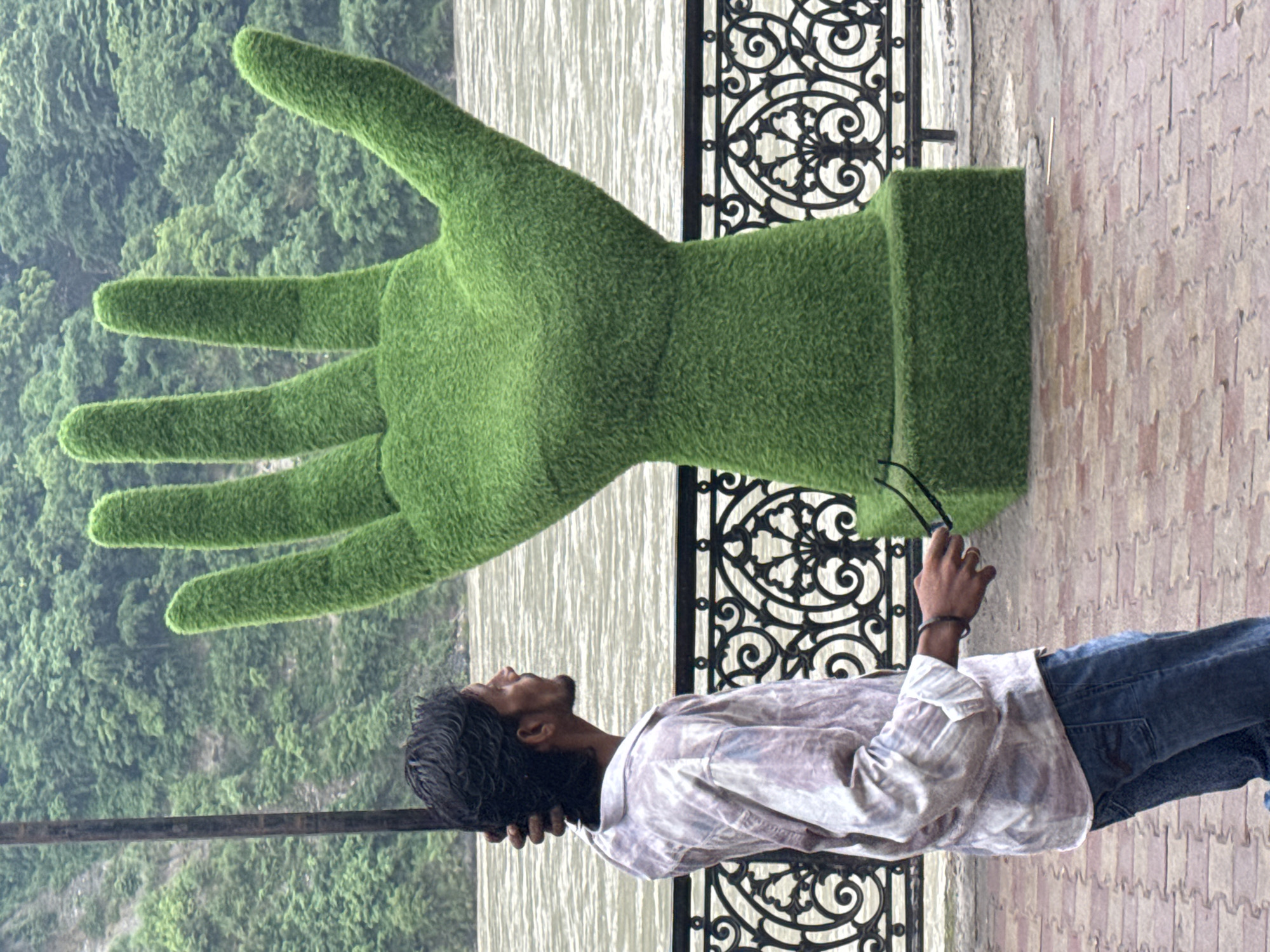A man is standing in front of a large, green, hand-shaped sculpture in Rishikesh, India. The sculpture is covered in artificial grass and is the main subject of the image. The man is facing to the right, looking at the sculpture. He is wearing a light-colored, patterned shirt and blue jeans. He is holding a pair of sunglasses in his right hand and has his left hand on the back of his head. In the background, a river can be seen, with lush green hills on the opposite bank. A decorative black metal fence runs along the walkway in front of the river. The walkway is paved with reddish-brown bricks.